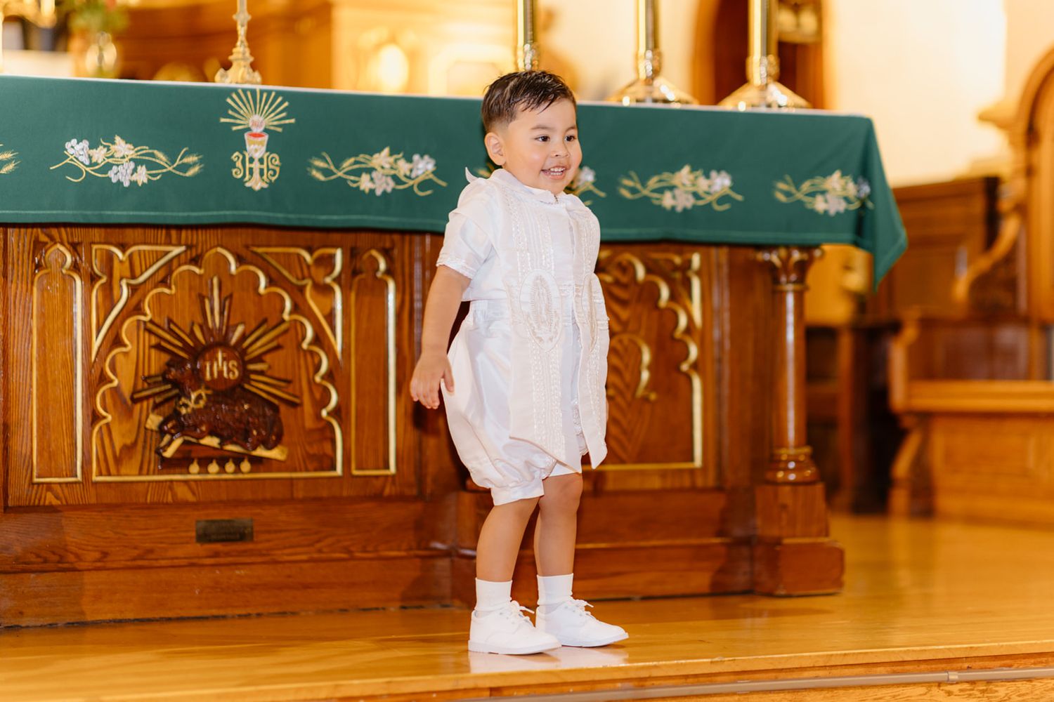 A smiling toddler in a white romper and socks stands on a wooden church floor near an ornate altar.