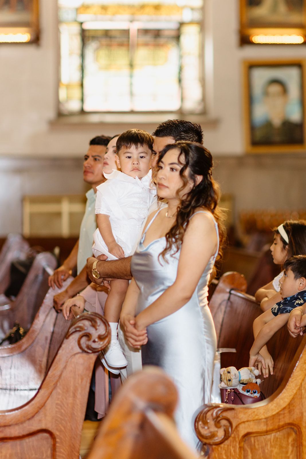 Mother in silver dress holds her toddler in white baptism outfit while standing in church pews.