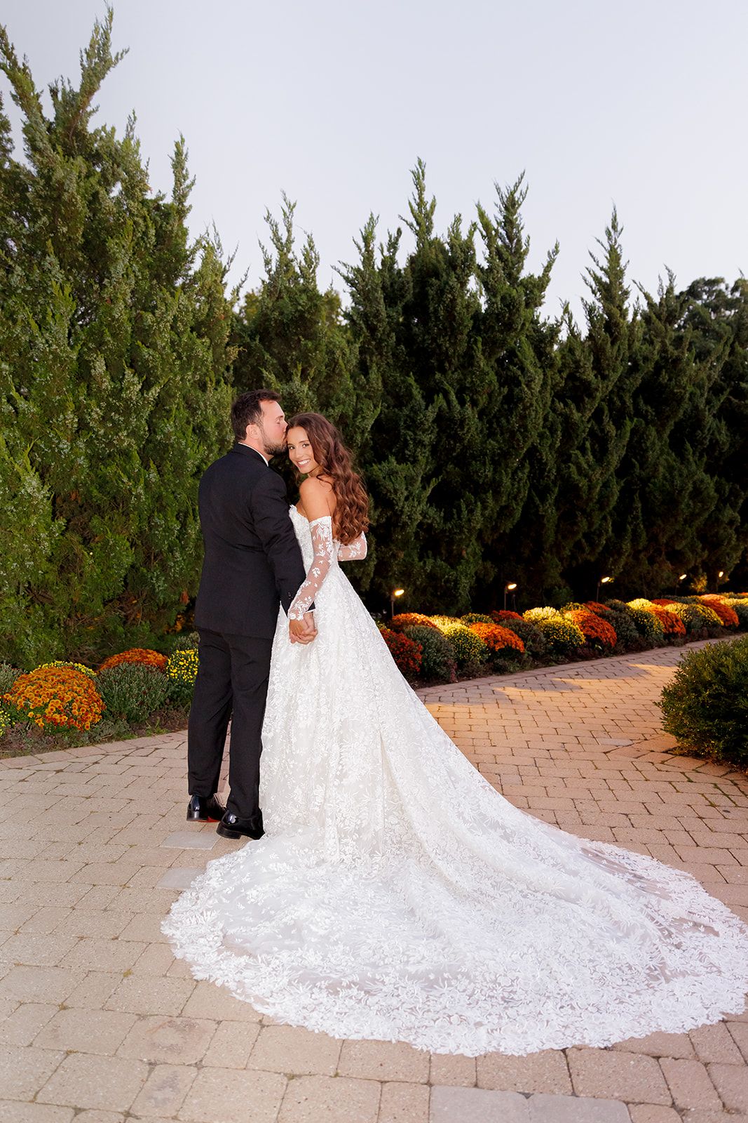 Bride in lace gown with long train embraces groom in black tuxedo outdoors among evergreen trees and colorful flowers.
