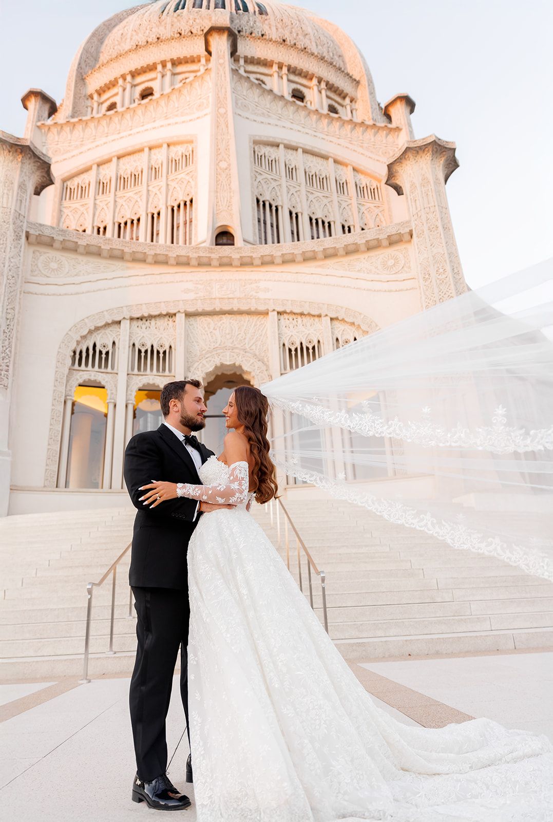 Bride and groom embrace before an ornate domed temple with veil flowing in the wind.
