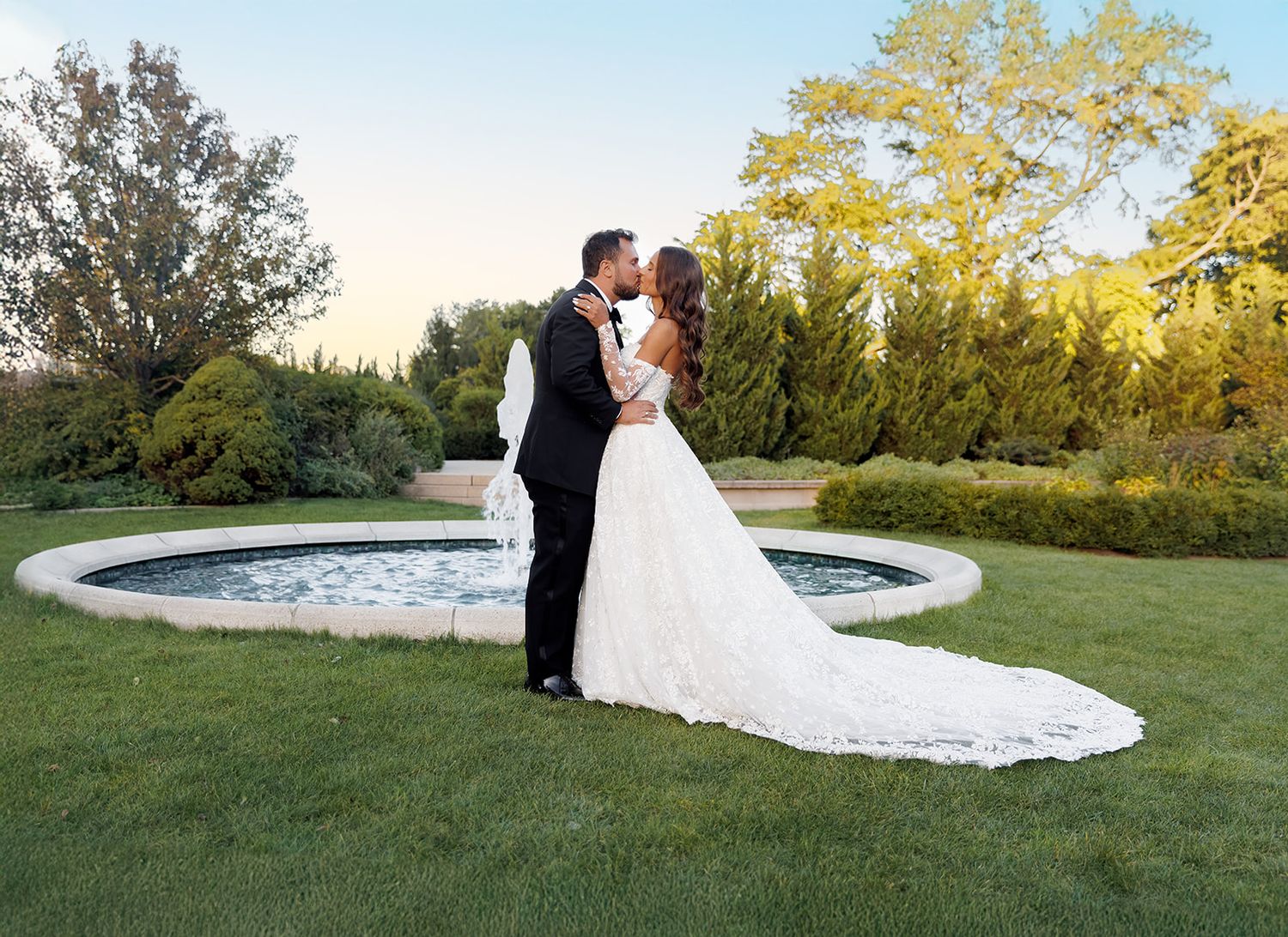 Newlyweds share a kiss on lush green lawn beside a circular fountain, framed by tall trees in golden hour light.