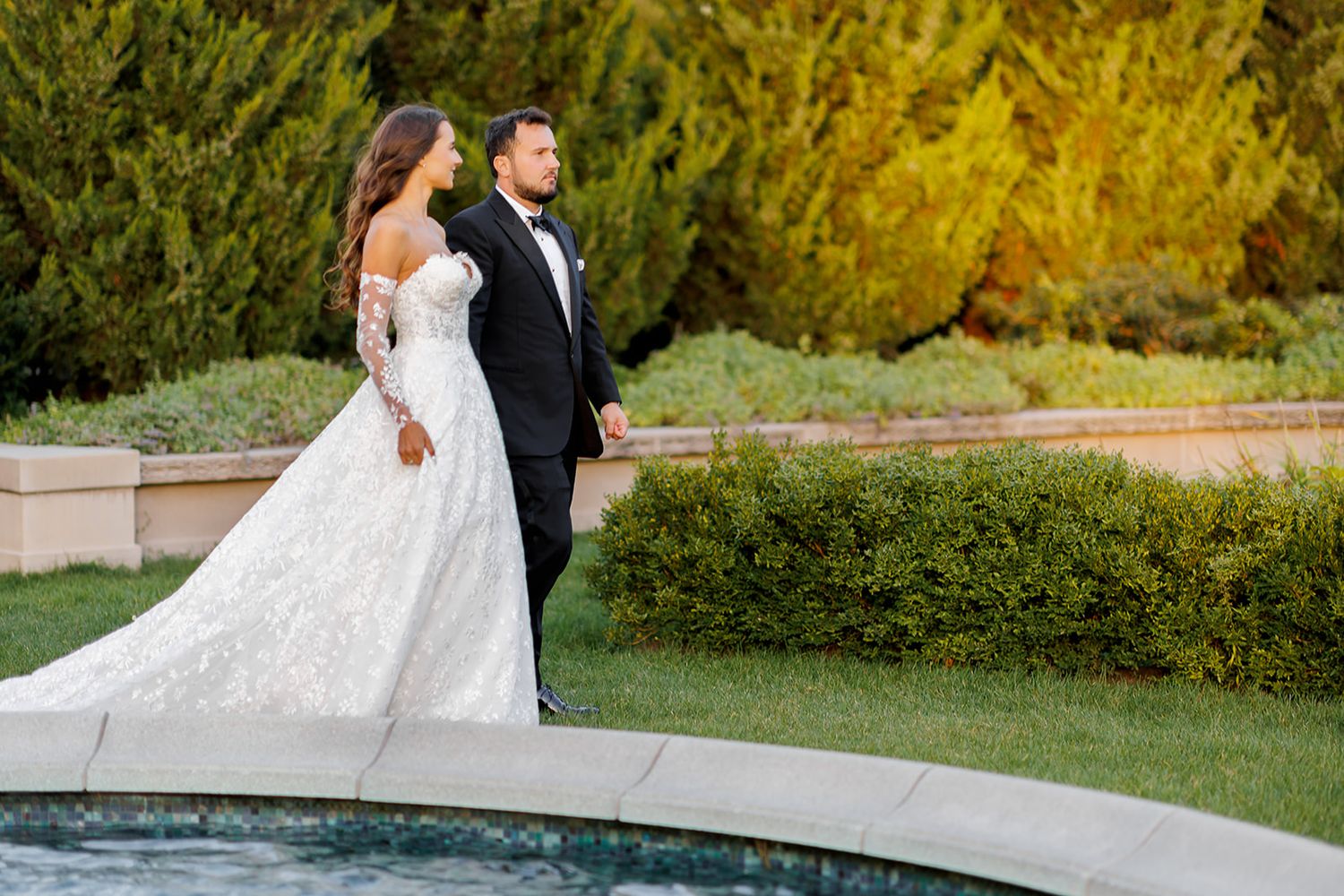 Elegant bride in lace gown and groom in tuxedo walk hand-in-hand beside a pool with autumn foliage.