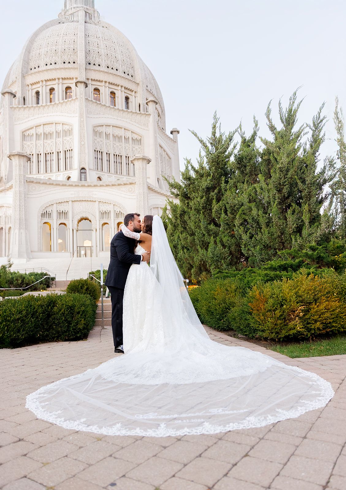 Newlyweds embrace and kiss before the ornate white Bahai Temple, bride wearing a sweeping lace-trimmed veil.