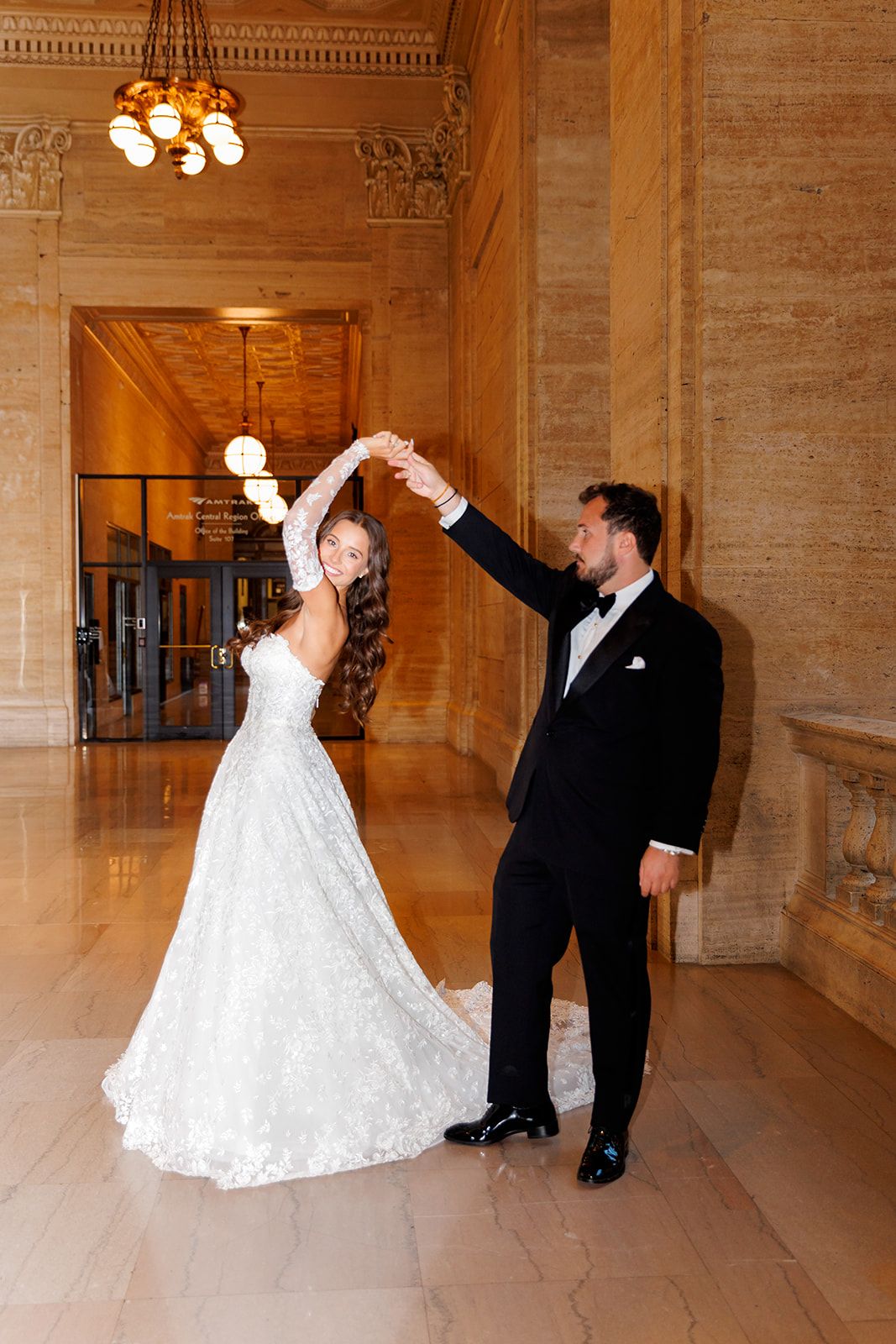 Bride and groom dancing elegantly in a grand marble hall, she in a lace gown, he in a black tuxedo.