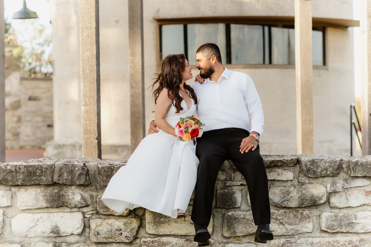 Couple in wedding attire share a romantic moment sitting on a stone wall outside a historic building.