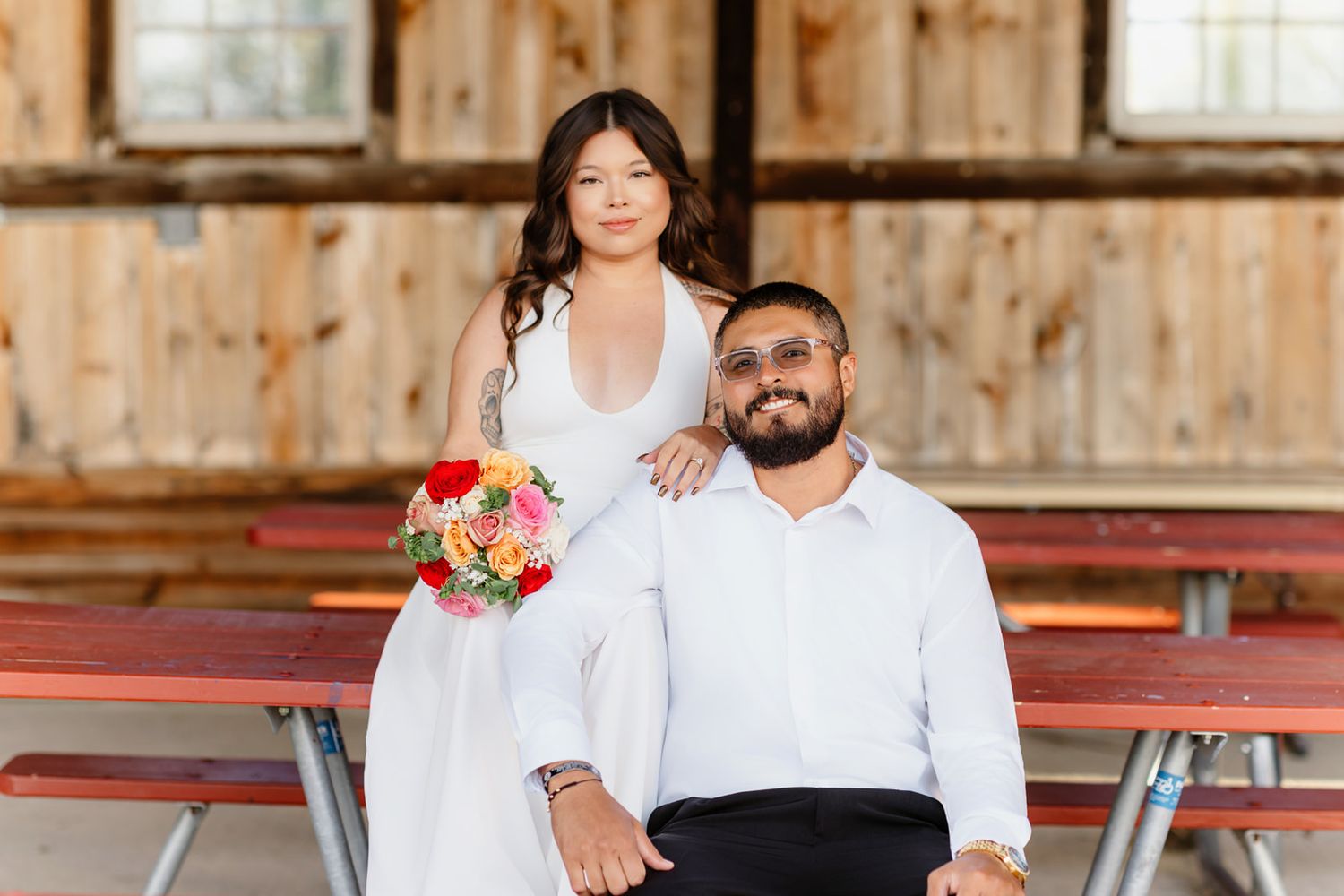 Bride in white gown holding colorful bouquet poses with groom in white shirt at rustic barn wedding venue.