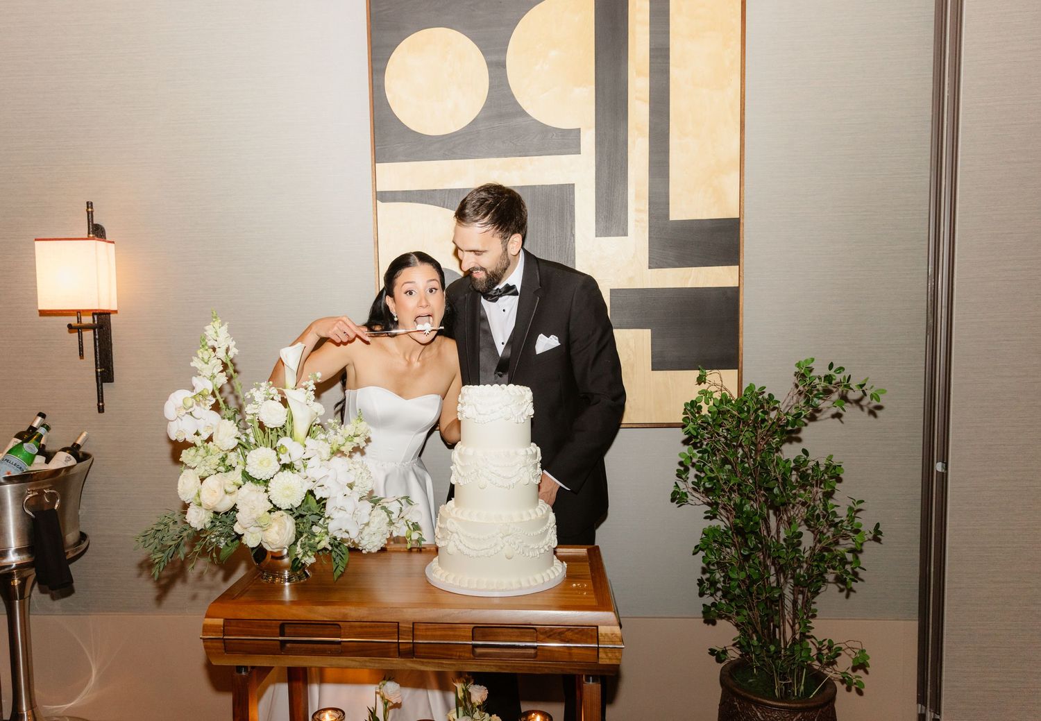 Bride and groom cut white tiered wedding cake beside white floral arrangement at elegant reception.