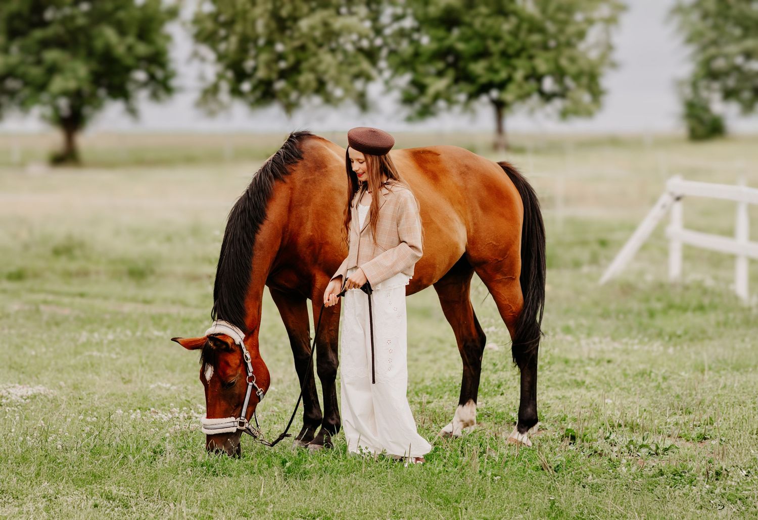 Woman in white dress and hat stands beside a grazing bay horse on a lush green pasture.