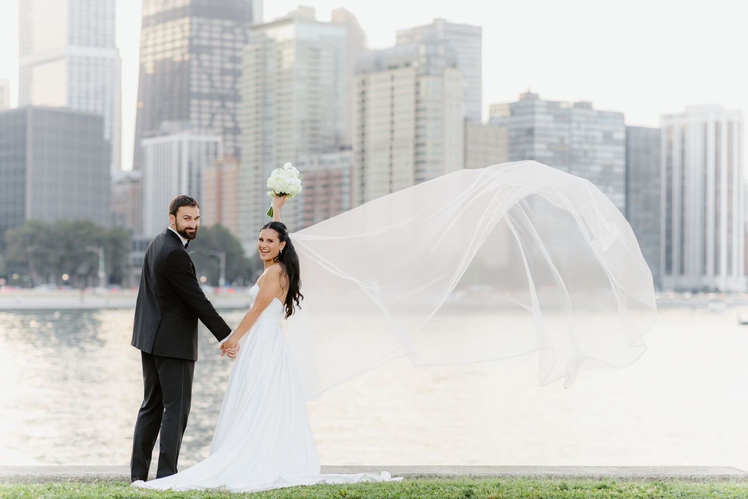 Bride and groom hold hands by Chicago lakefront, her veil flowing dramatically in the wind with city skyline behind.