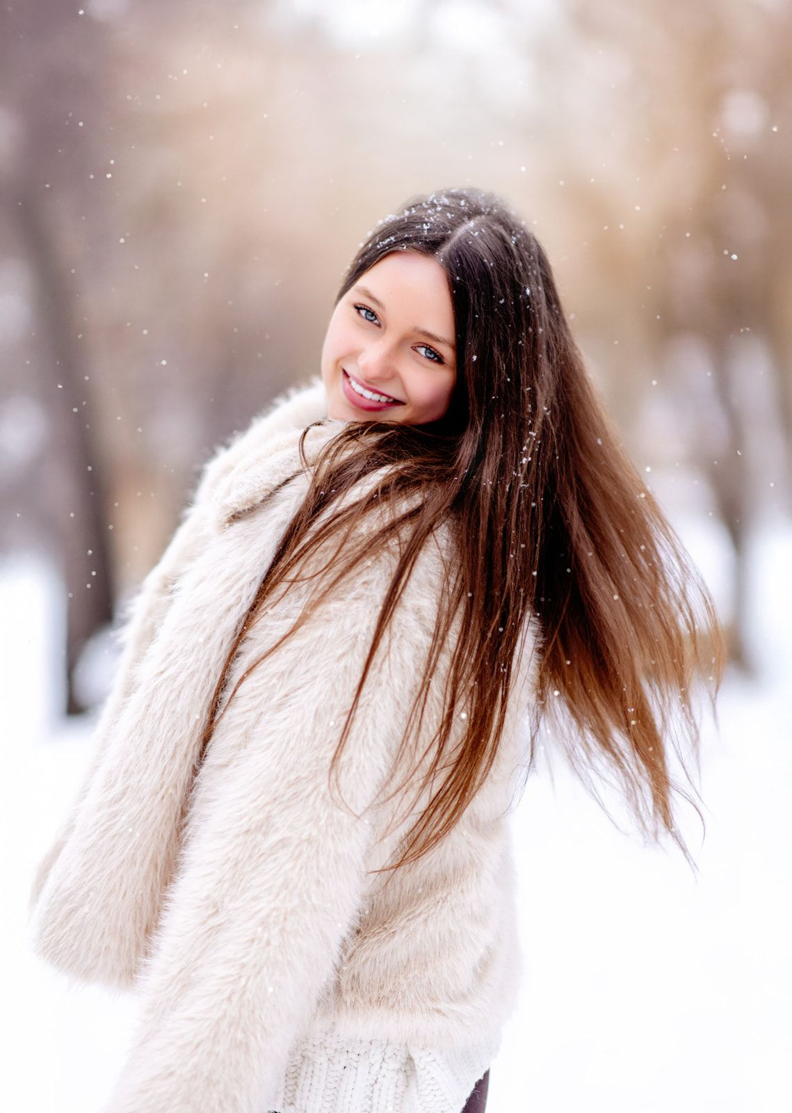 Smiling young woman in white fluffy coat with windswept hair enjoying snowfall in winter park.