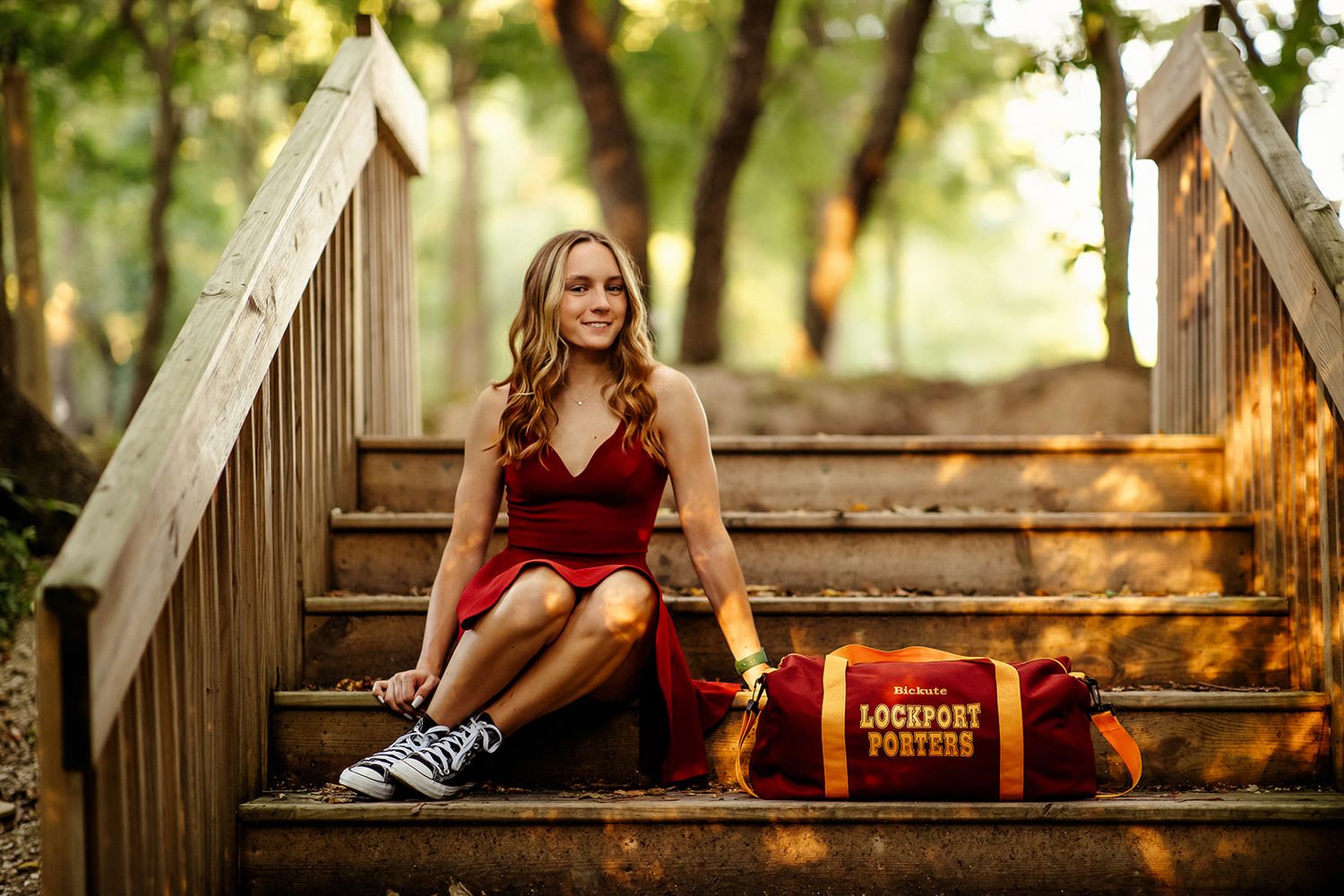 Smiling young woman in red dress sits on wooden stairs with a Lockport Porters sports bag.