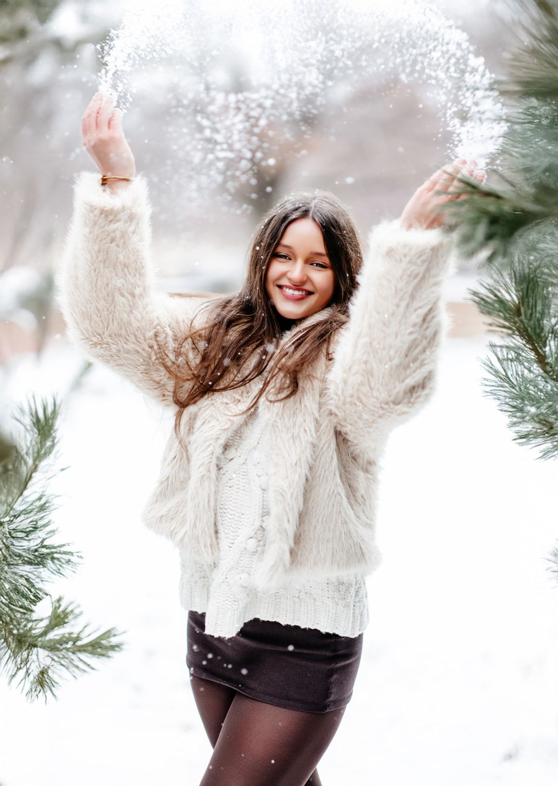 Young woman in white fluffy jacket tosses snow joyfully outdoors among snow-dusted pine trees in winter.