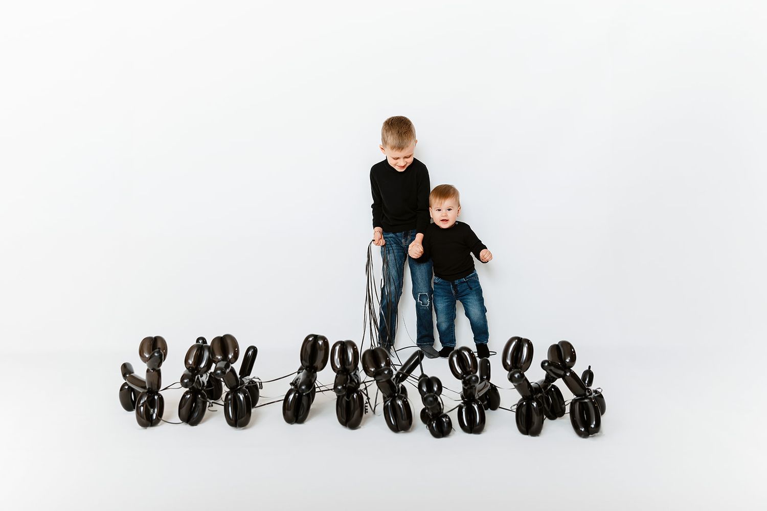 Two young boys in black shirts pose with multiple shiny black balloon animal dogs on a white background.