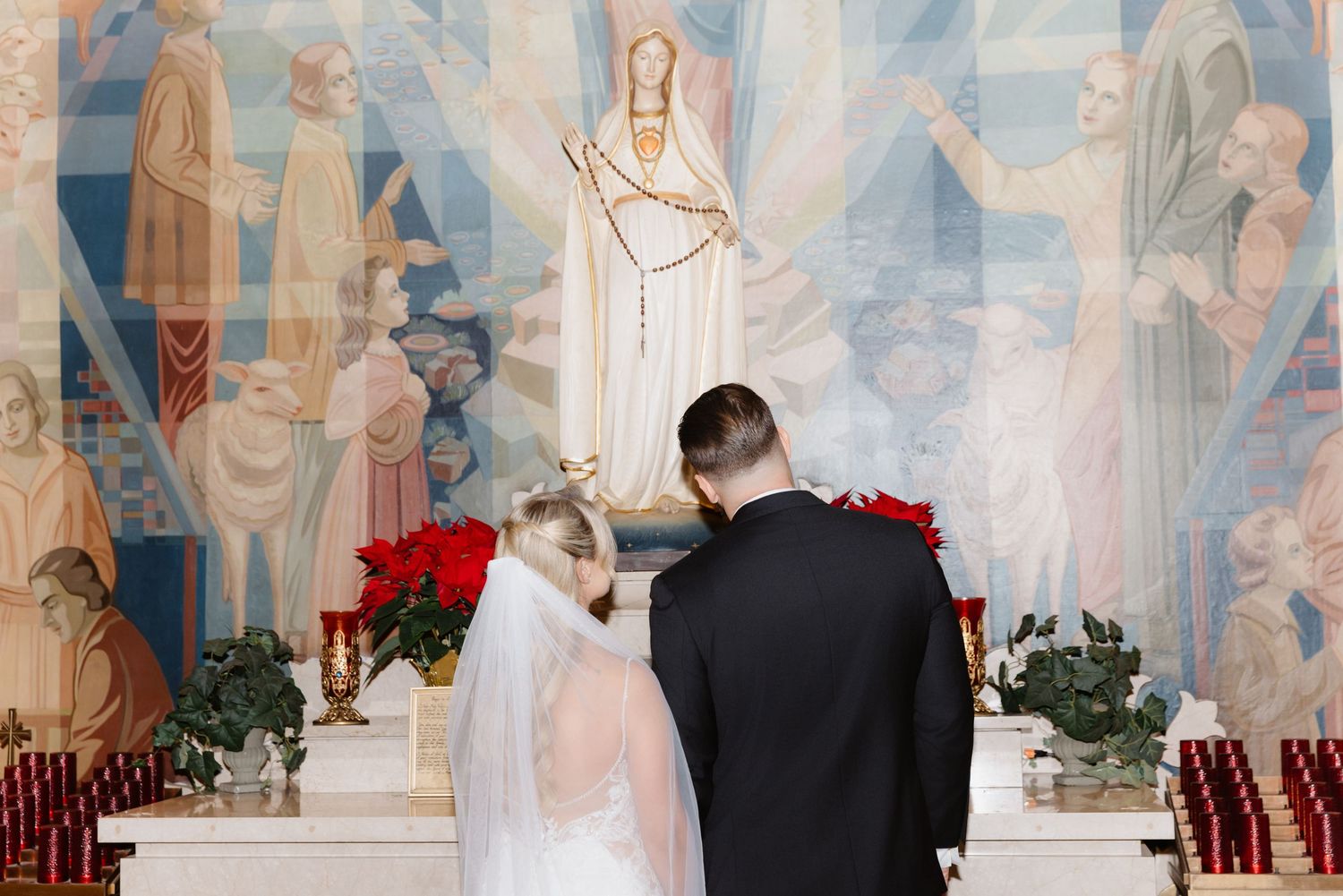 Bride and groom standing at ornate altar before a colorful religious mural with red roses and candles.
