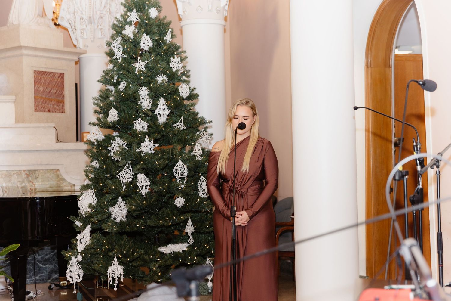 Woman in brown dress singing near decorated Christmas tree at wedding venue.