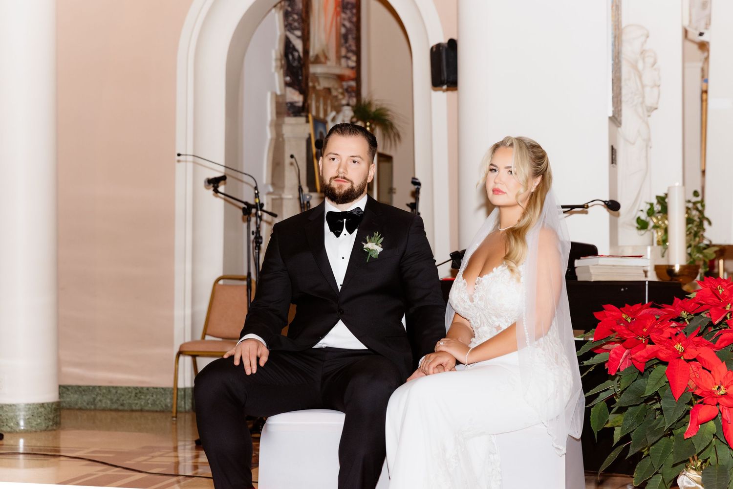 Close-up of bride and groom seated at church altar with red poinsettias in background.