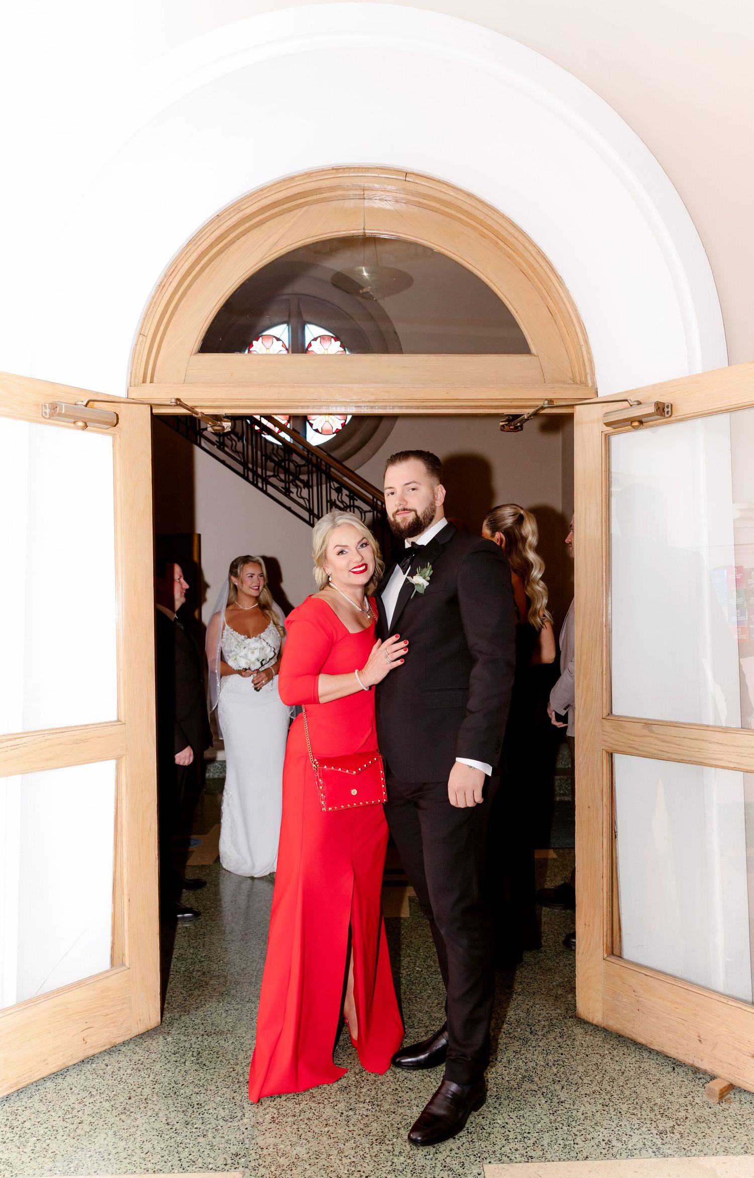 Groom in black tuxedo poses with woman in red gown at church entrance during wedding ceremony.