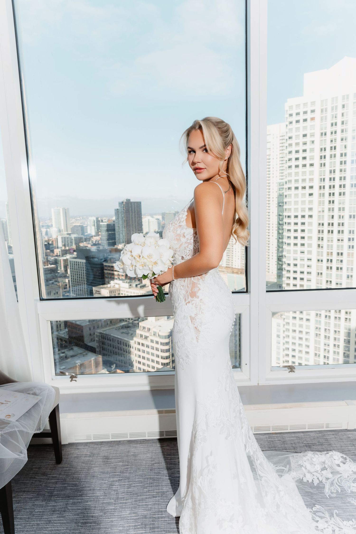 Blonde bride in lace wedding dress holds white bouquet while posing before sunny city skyline.