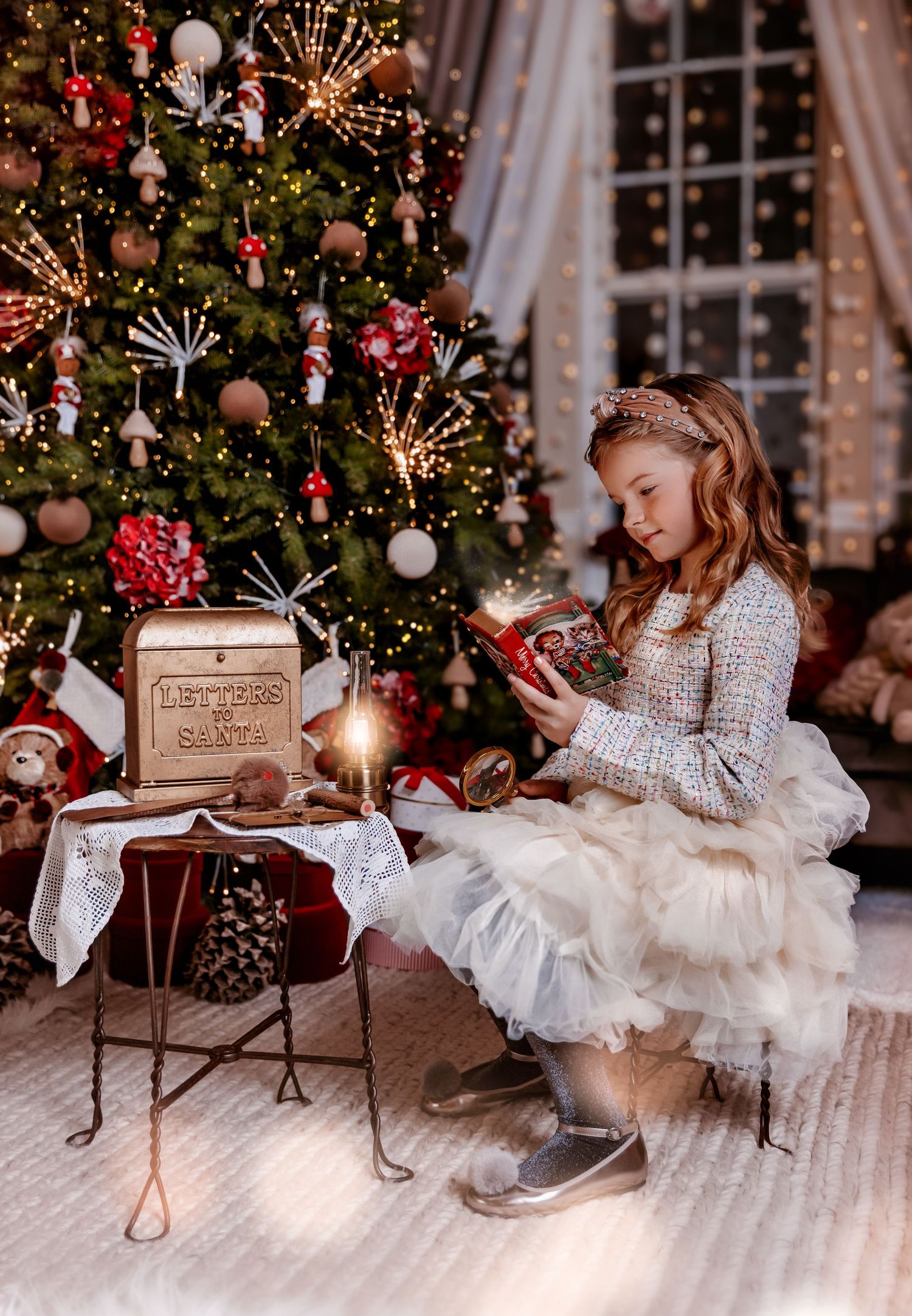 Girl in white dress reads book by Letters to Santa mailbox near decorated Christmas tree with warm lights.