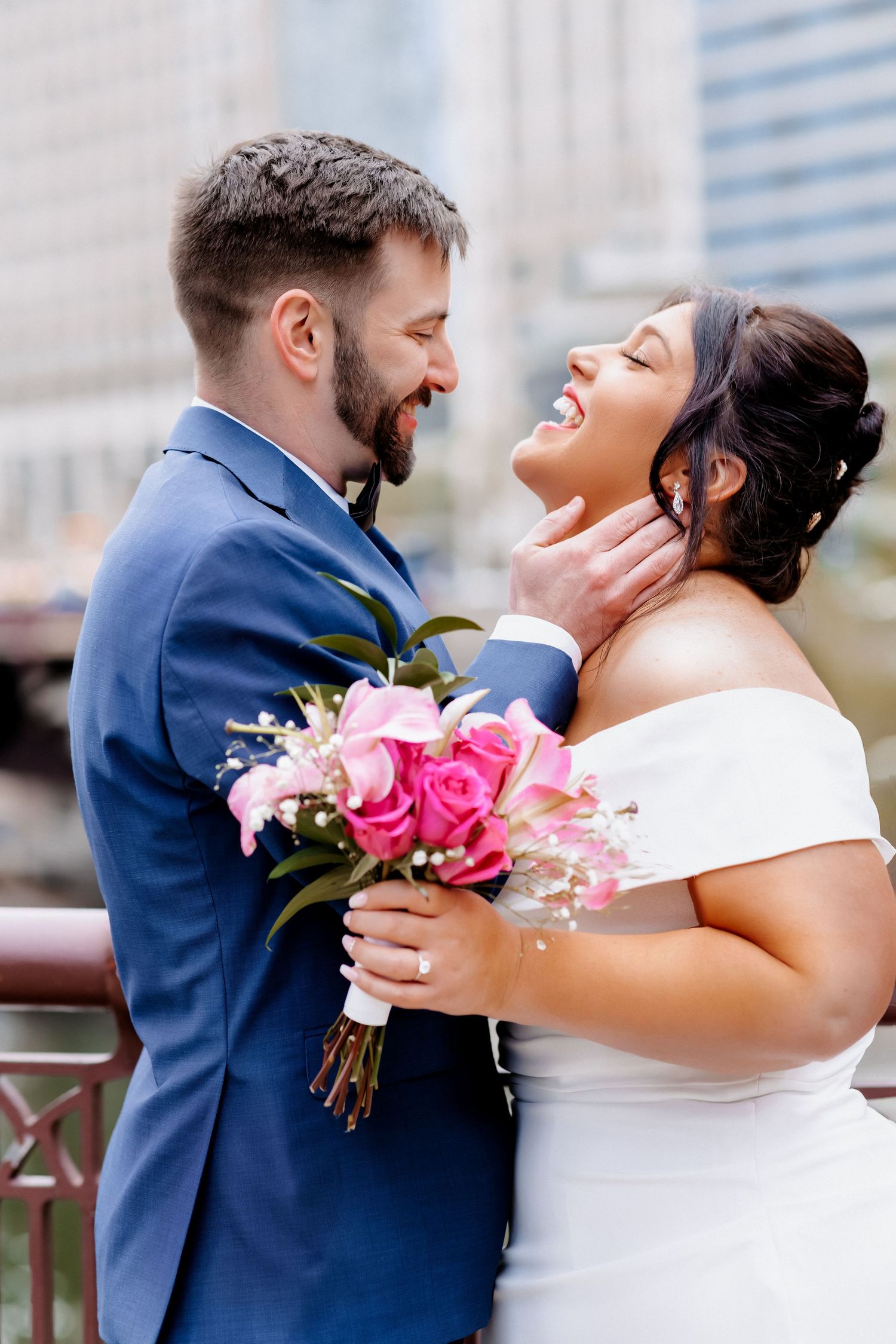 Laughing bride in white gown and bearded groom in blue suit share joyful moment on city bridge with pink bouquet.