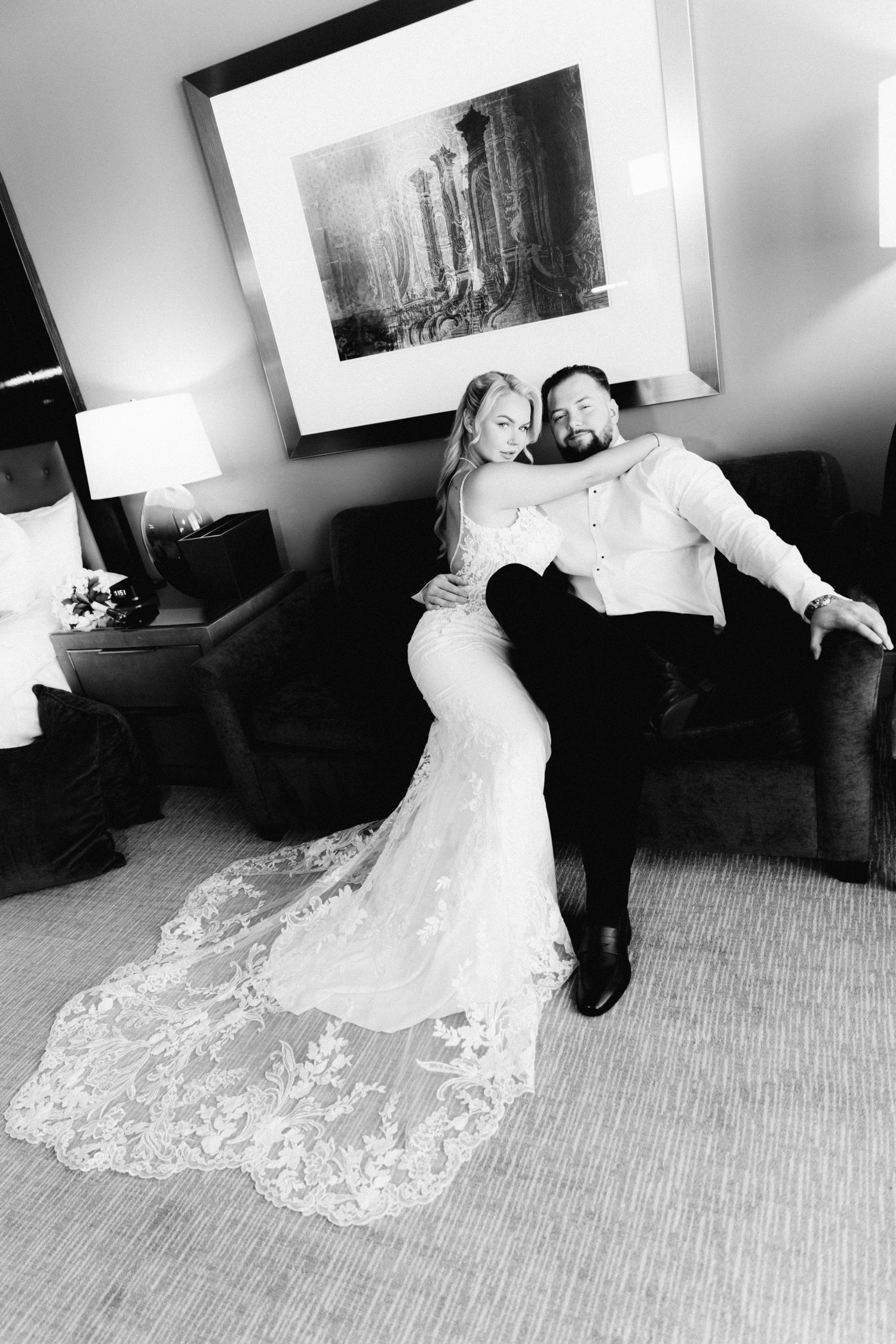 Black and white photo of bride in lace gown and groom in white shirt sitting together on sofa.