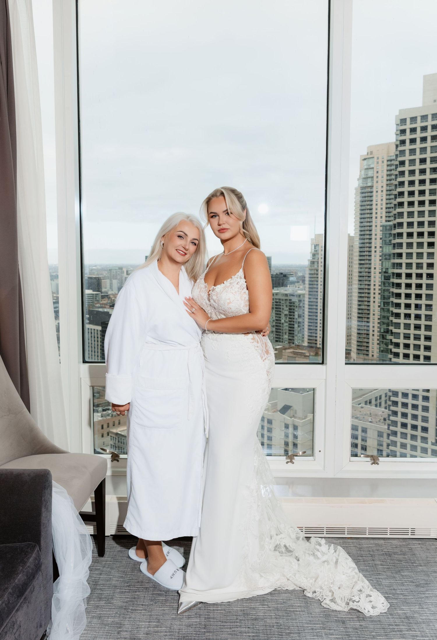 Bride in fitted lace wedding dress poses with older woman in white robe by city window.