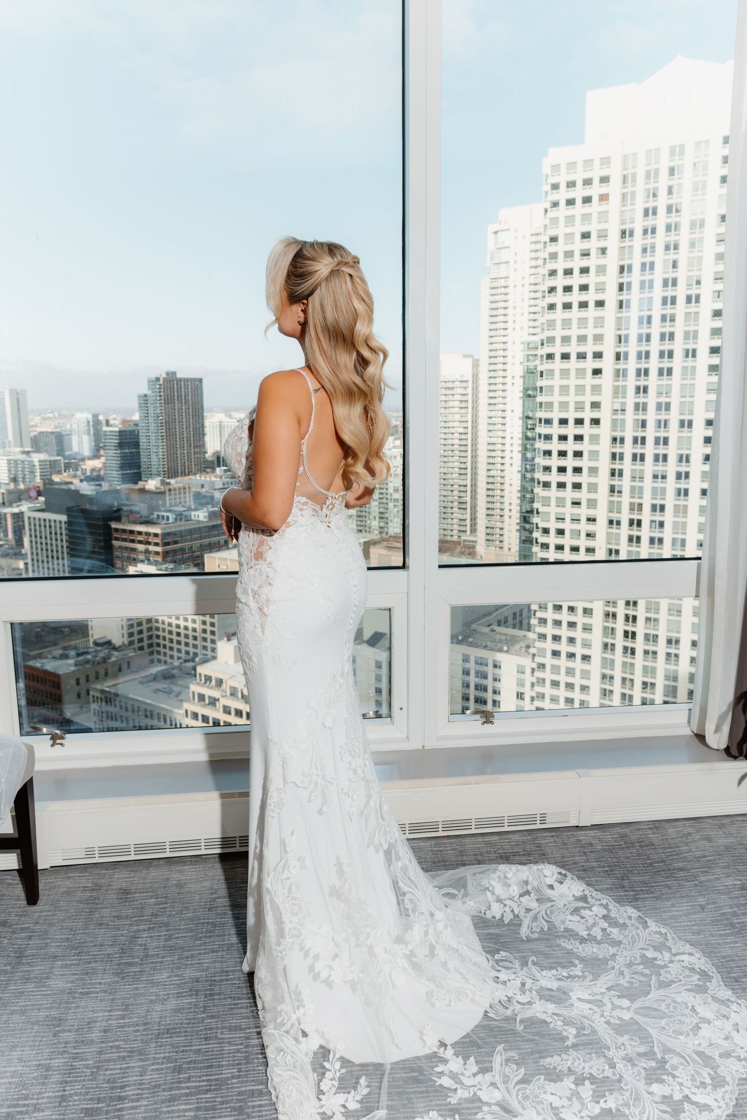 Bride in fitted lace wedding gown with long train gazes at city skyline through large windows.