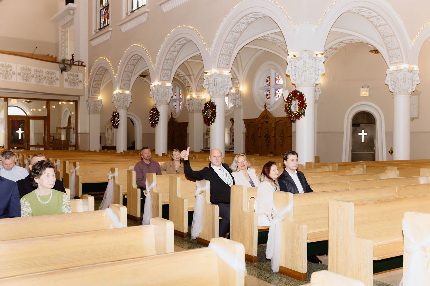 Guests seated in elegant white church pews during a wedding ceremony with arched ceilings and holiday wreaths.