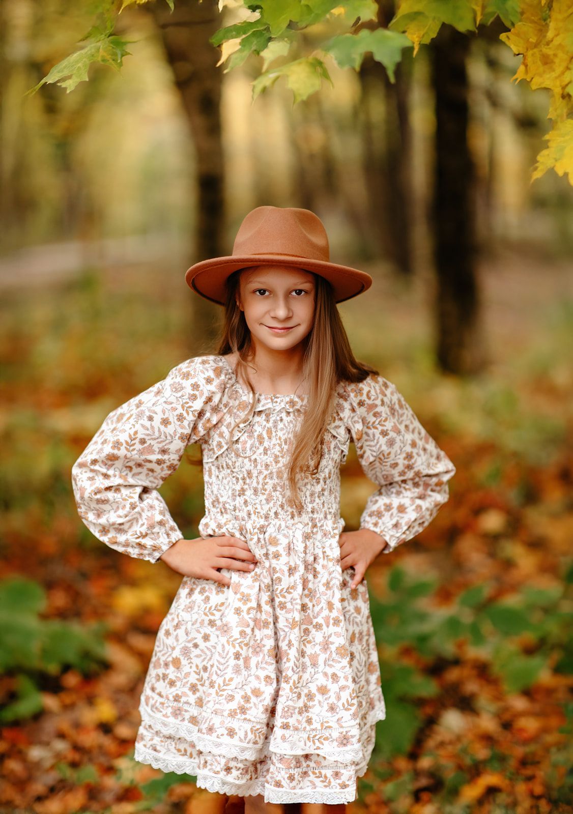 Young girl in floral dress and brown hat poses in autumn forest with colorful fallen leaves.