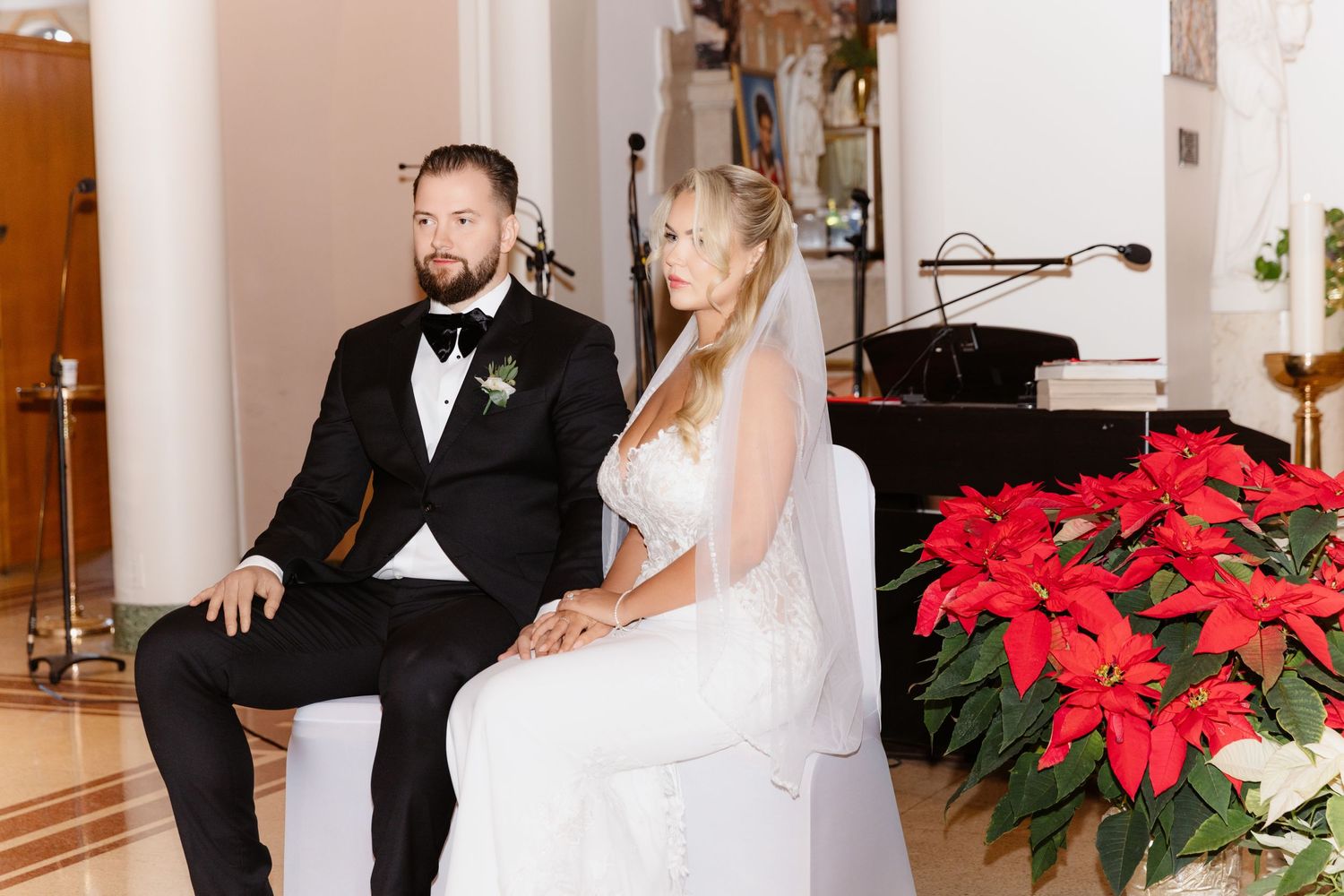 Bride in lace gown and groom in black tuxedo sit attentively during ceremony beside vibrant red poinsettias.