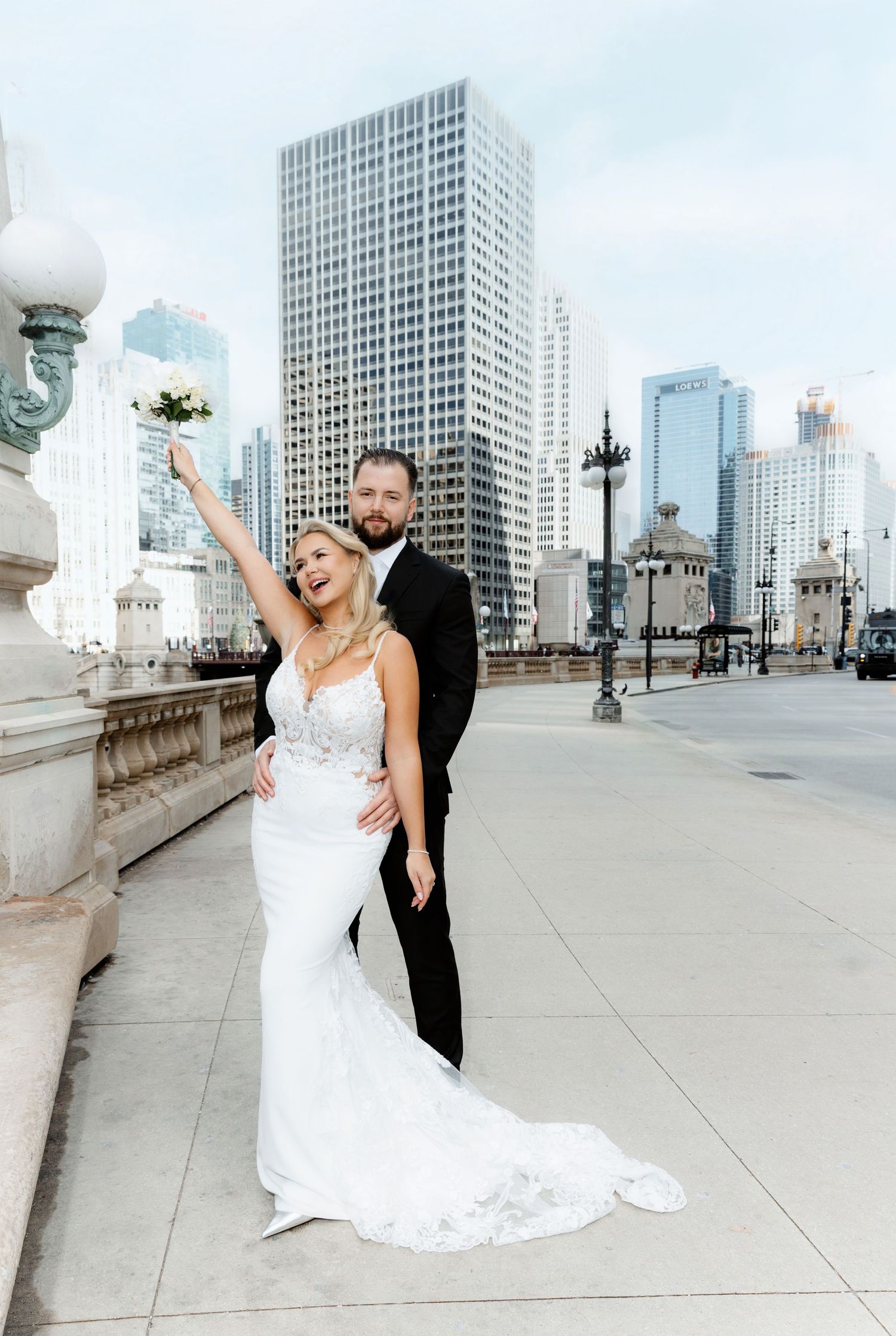 Bride in white gown raises bouquet as groom holds her close on Chicago bridge with city skyline backdrop.