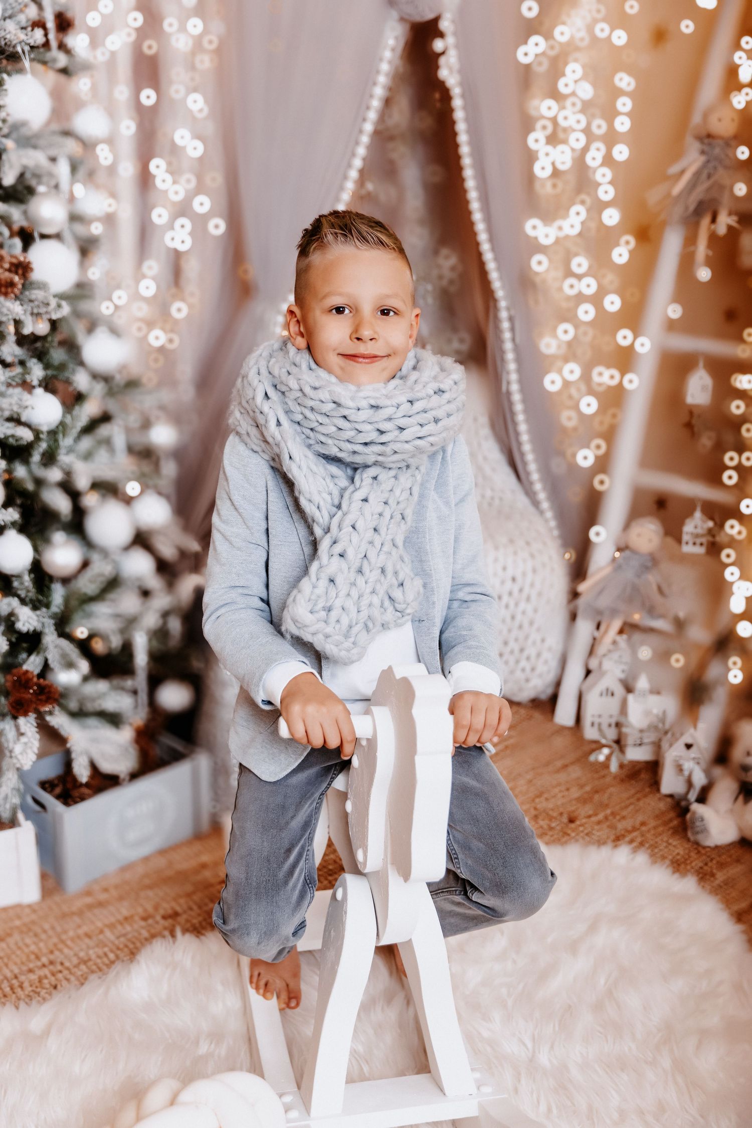 A smiling boy in a blue sweater and gray knit scarf sits on a white rocking horse in a festive Christmas decor.