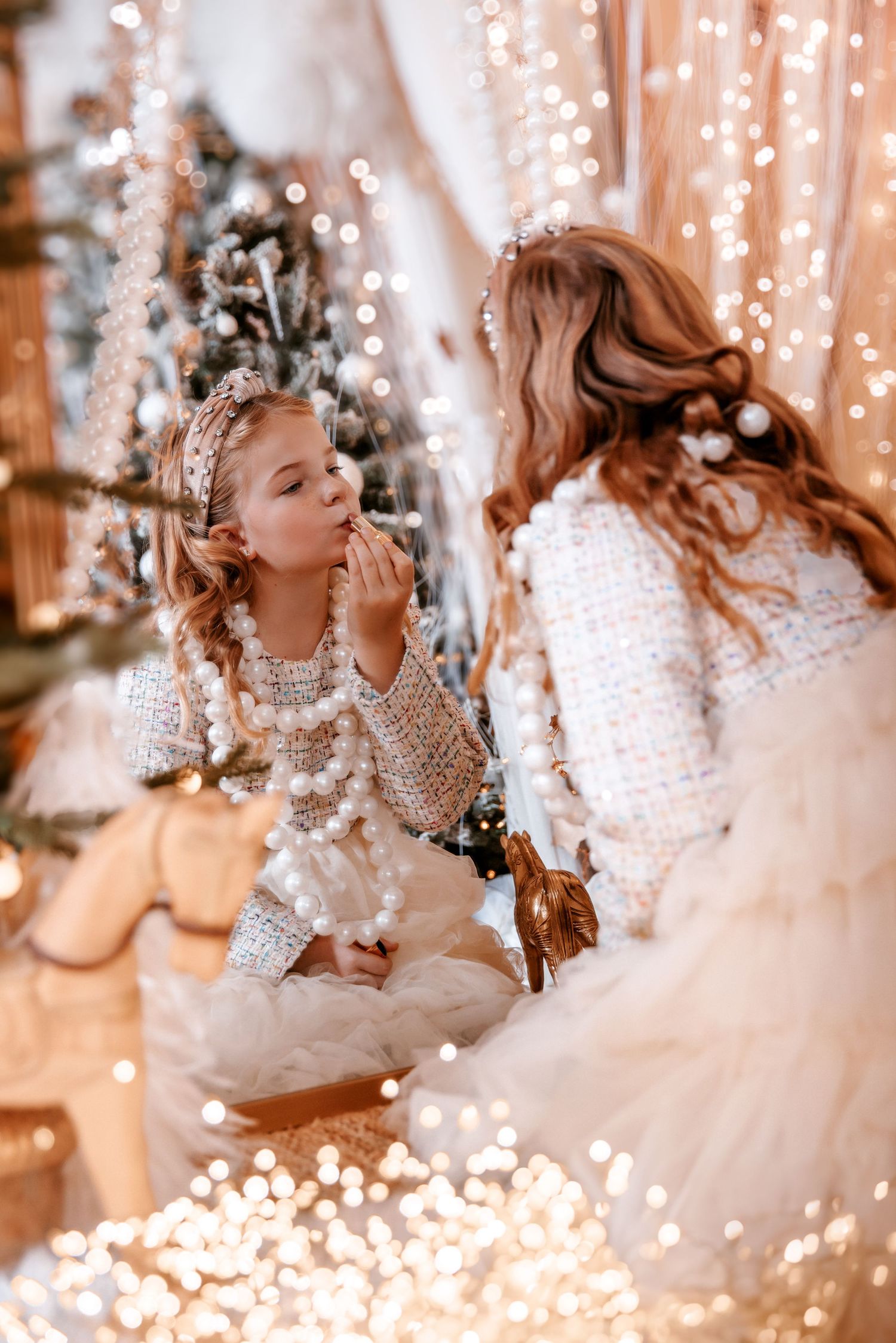 Young girl applies lip gloss while sitting near sparkling Christmas lights and holiday decor.