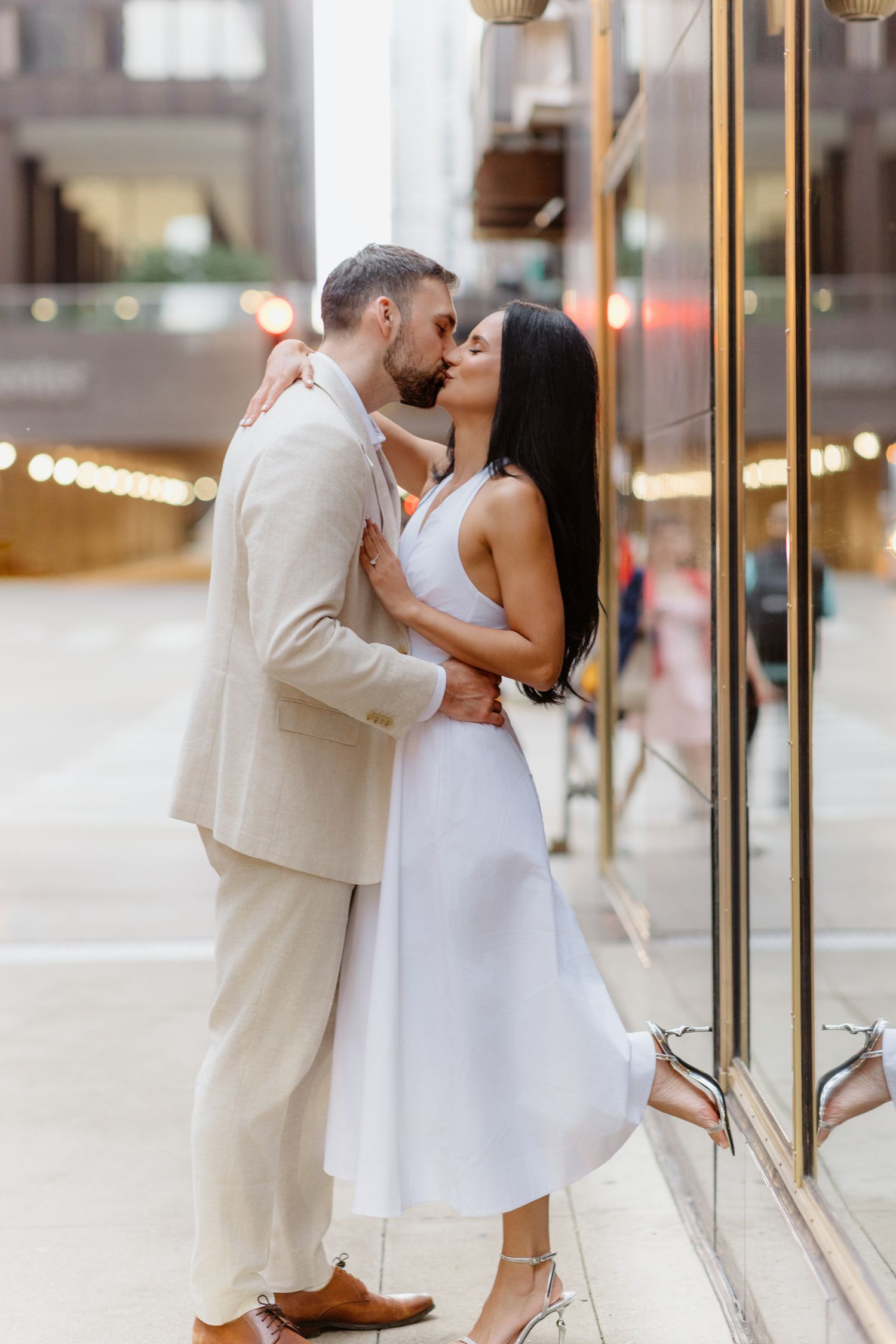 Couple sharing a passionate kiss beside a gold-trimmed glass building facade on a city street.