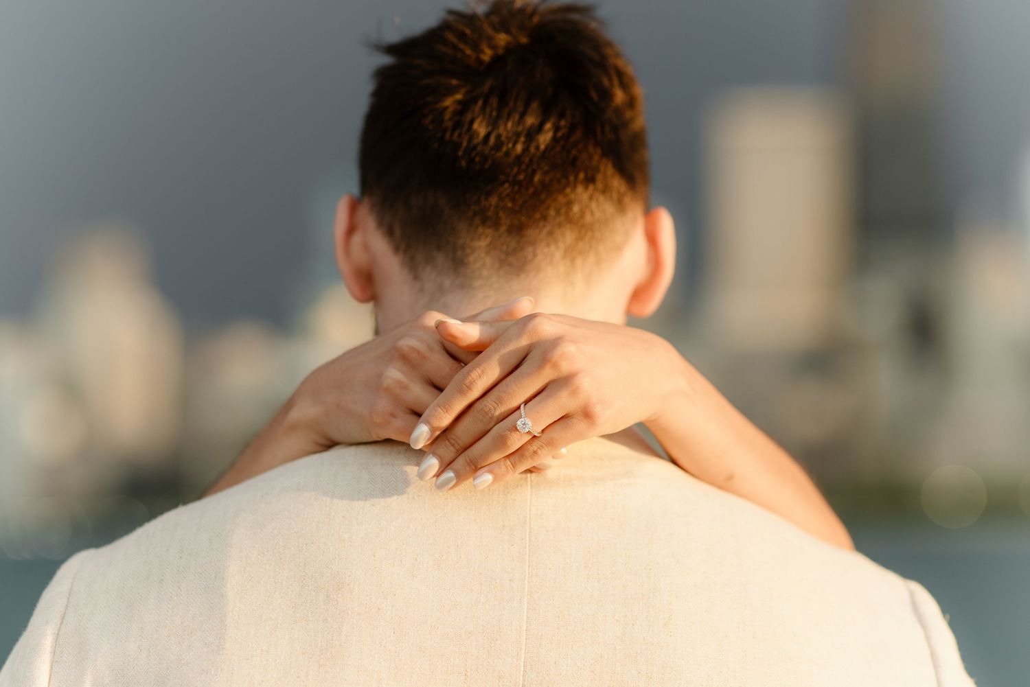 Close-up of womans hand with engagement ring resting on mans shoulder with blurred city skyline behind.