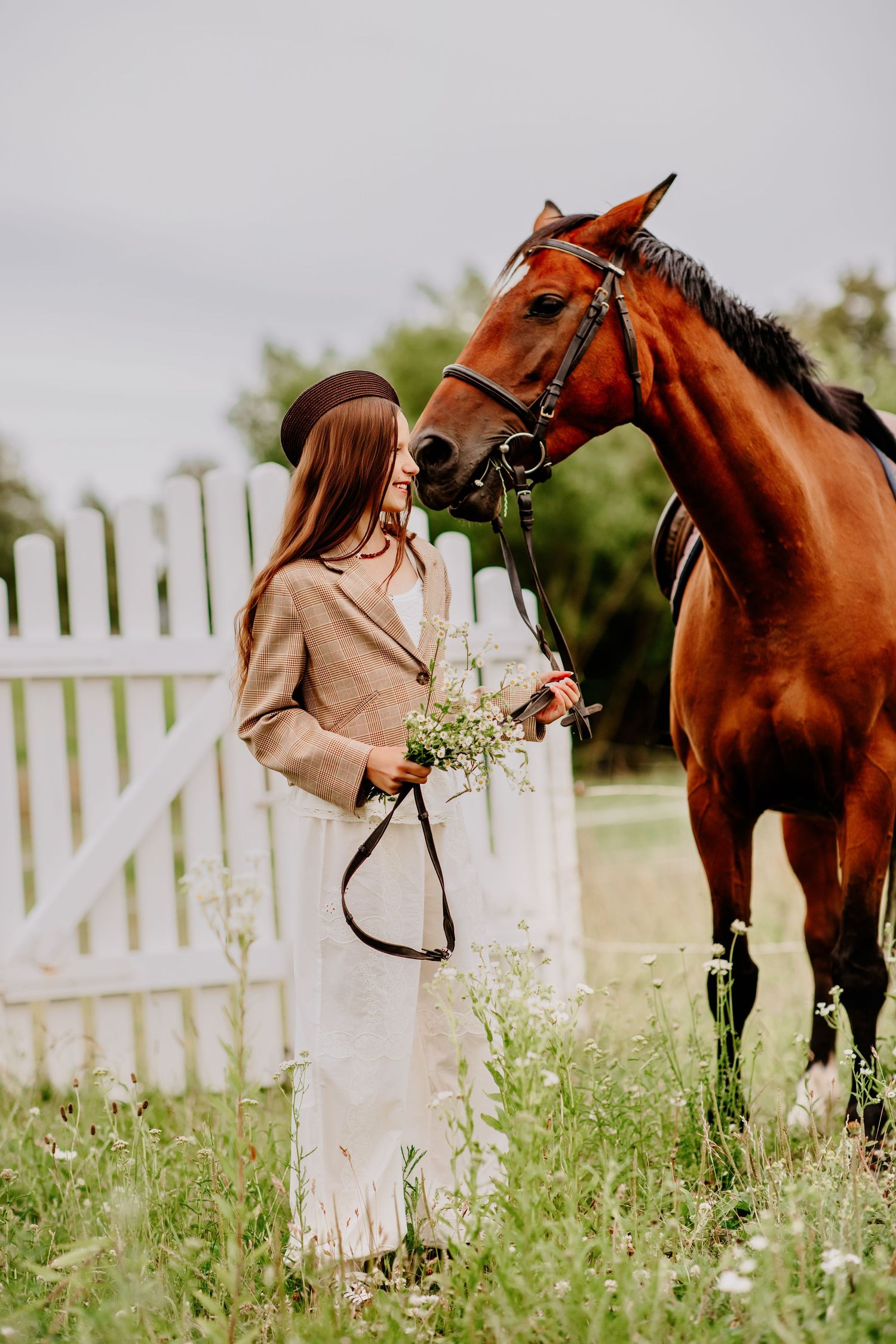 Young woman in brown hat and beige coat holds wildflowers while a bay horse nuzzles her near a white fence.