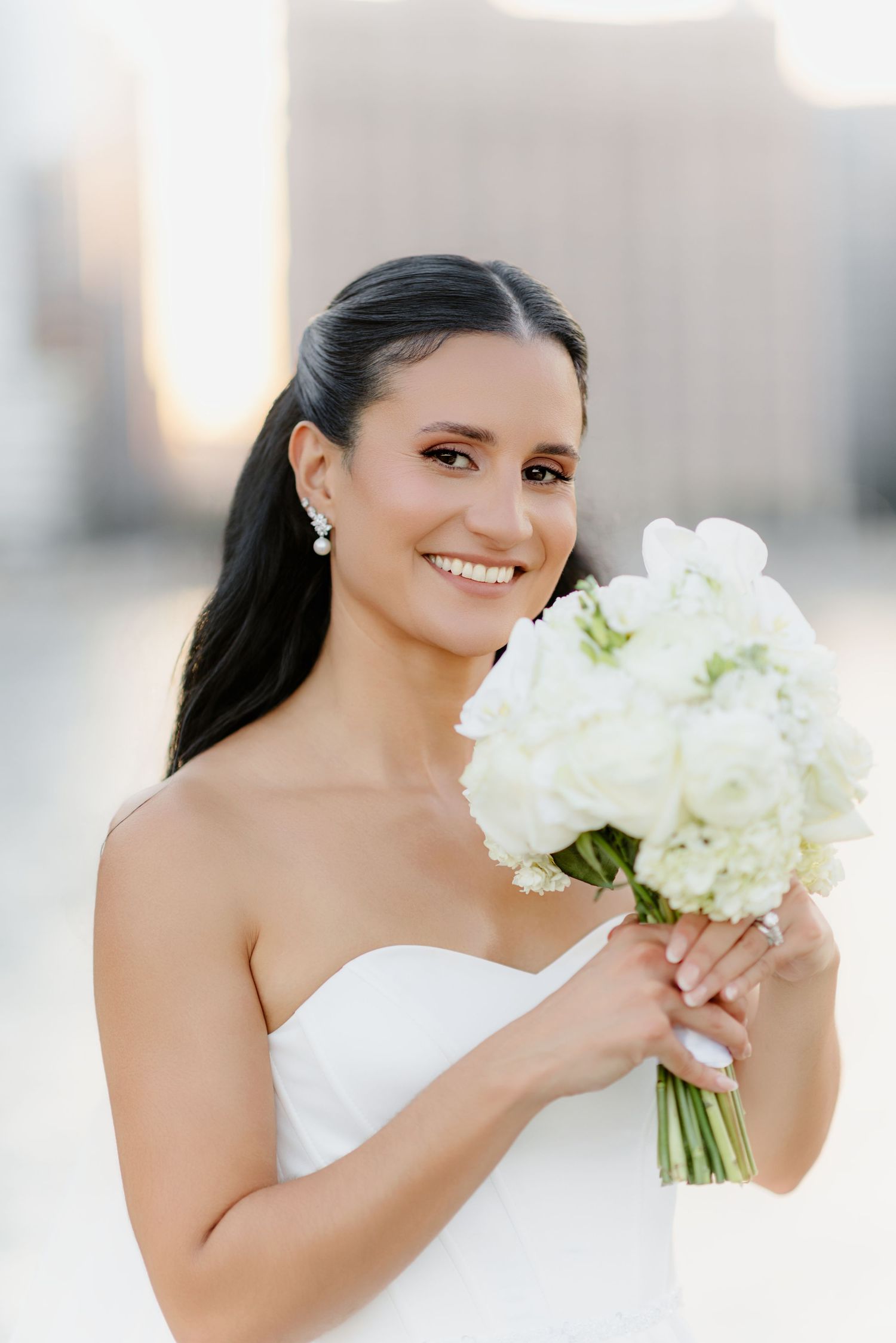 Smiling bride in strapless white gown holding elegant white floral bouquet with city skyline backdrop.
