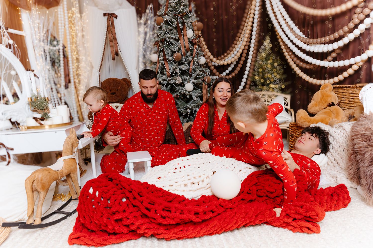 Family in matching red Christmas pajamas gathered around a white knit blanket near a decorated holiday tree.