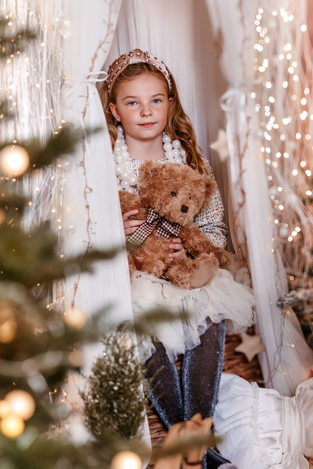 Young girl with tiara holding teddy bear amid Christmas lights and white canopy decorations in festive holiday setting.