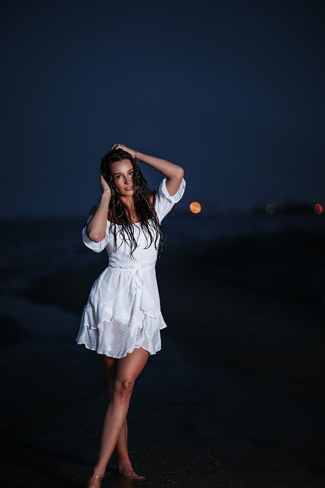 Brunette woman in white dress stands on dark beach at night with city lights behind her.