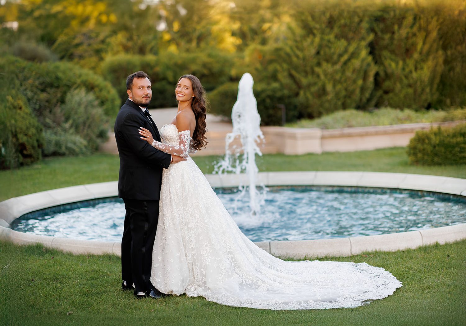 Newlyweds pose romantically by garden fountain, bride in lace gown with long train at sunset.