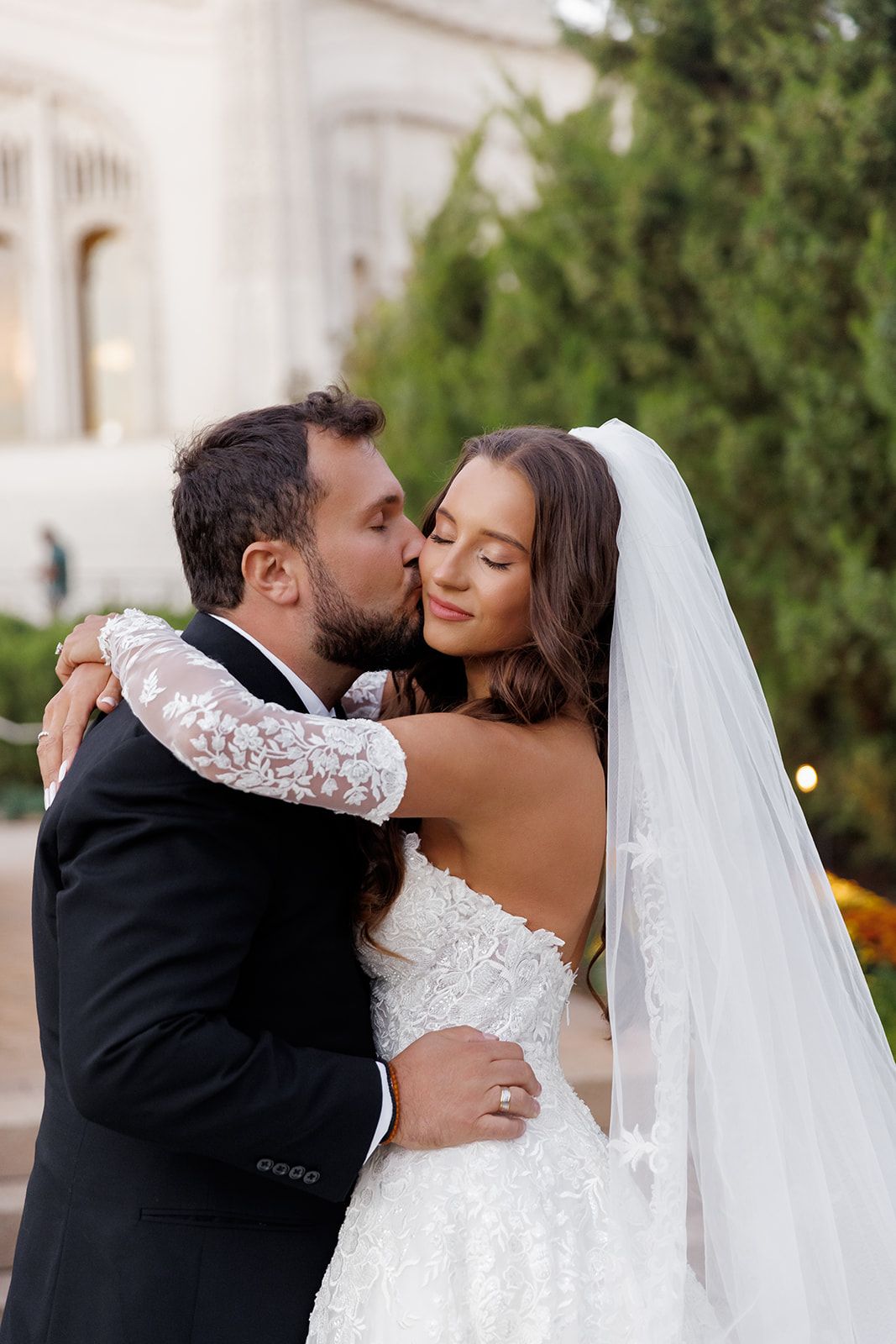 Groom in black tuxedo kissing bride on cheek outdoors, bride in lace wedding gown with veil.
