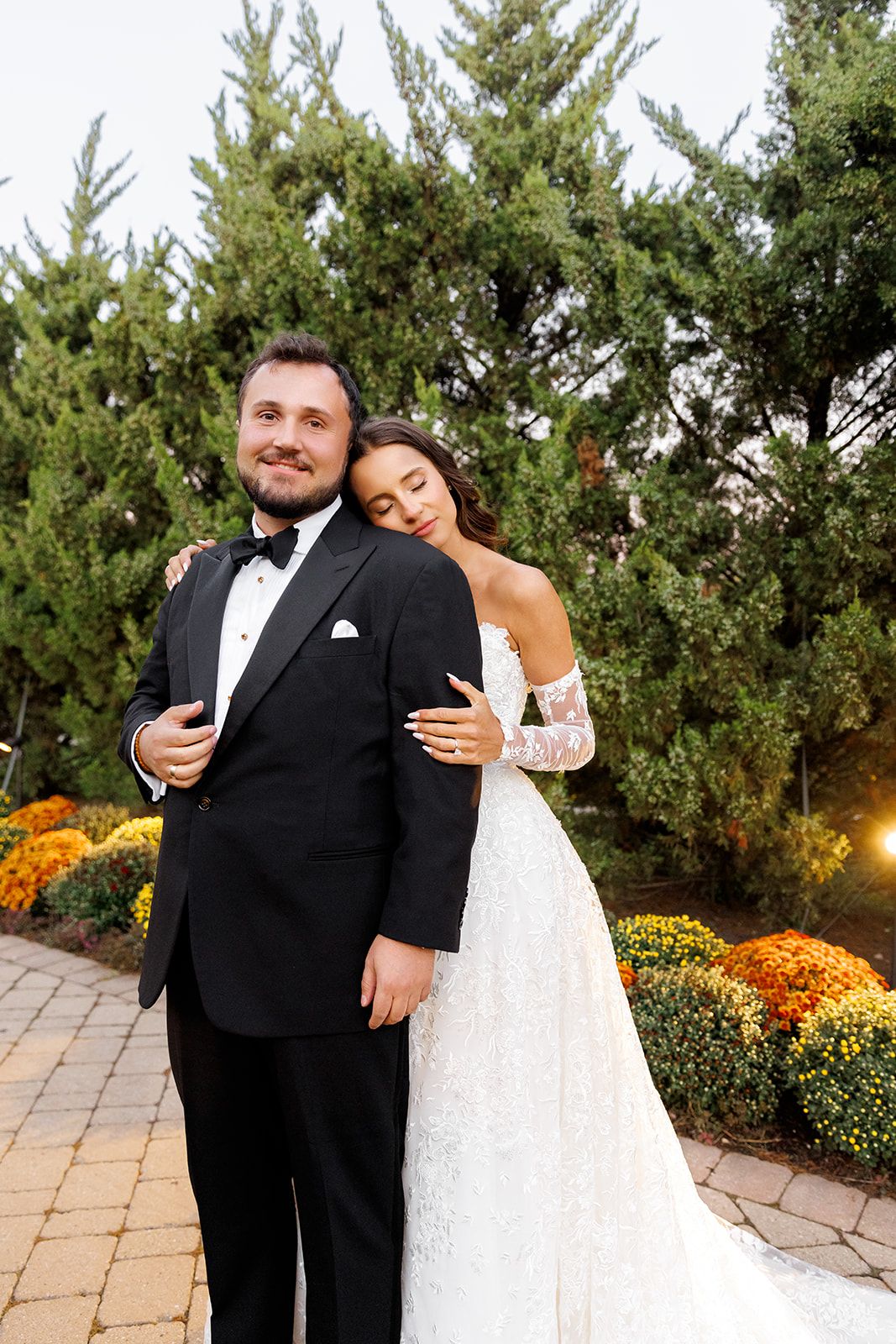 A bride in a lace gown embraces her groom in a black tuxedo outdoors, surrounded by evergreens and autumn flowers.