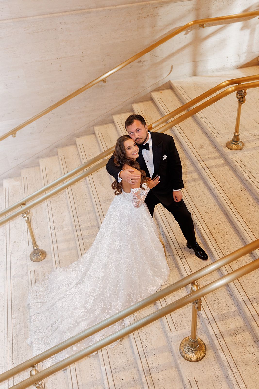 Bride and groom embracing on elegant marble staircase with gold railings, aerial view of wedding couple in formal attire.