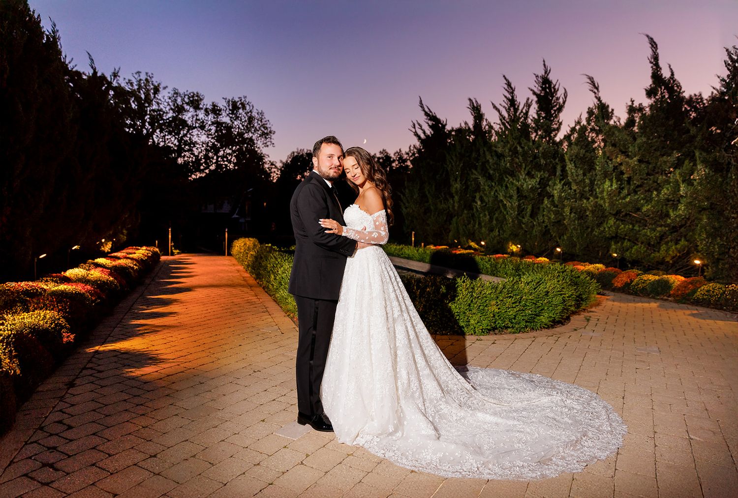 Bride and groom embrace on a garden patio at dusk, with warm pathway lights and tall hedges in the background.