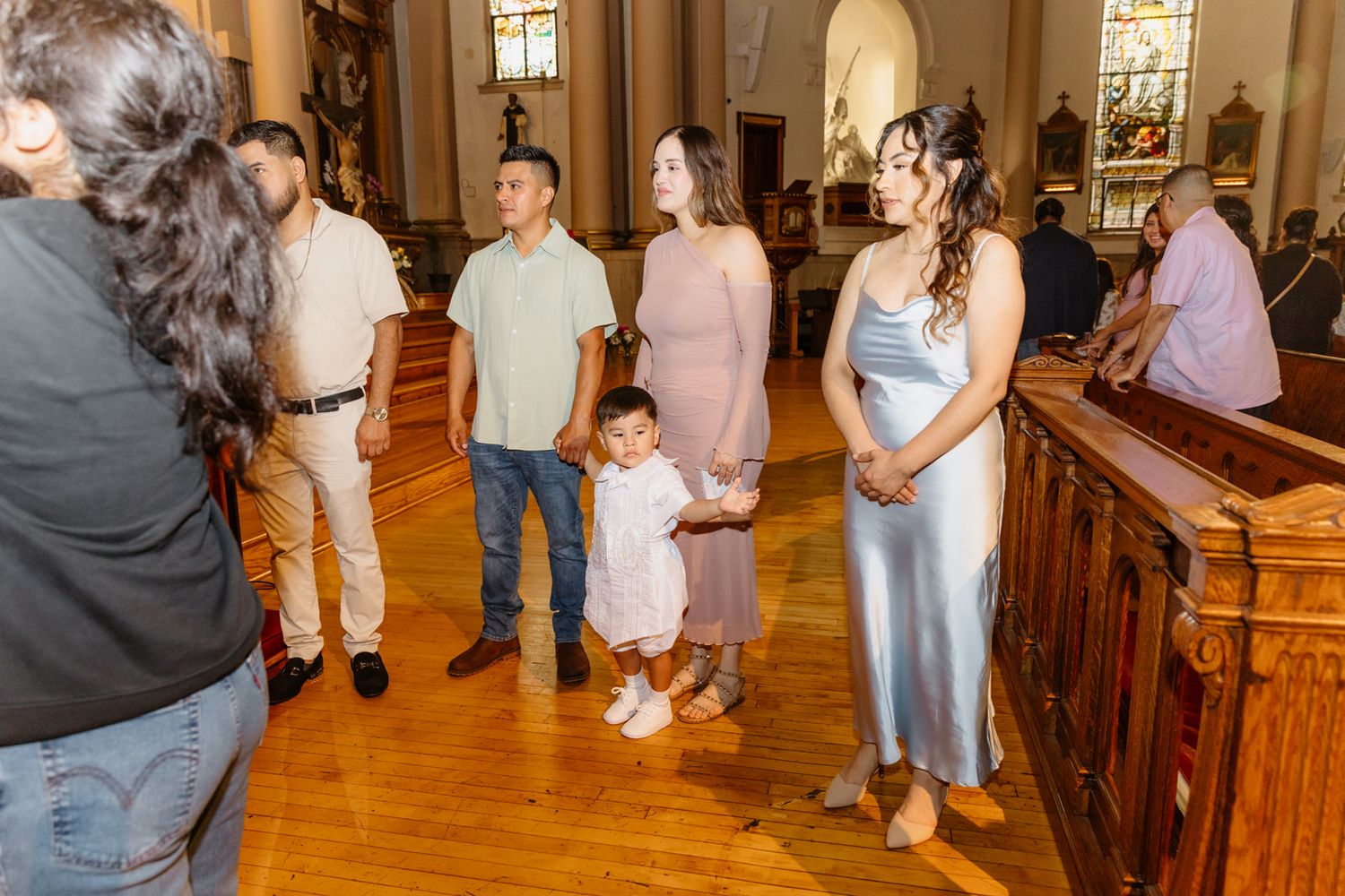 Family gathering in a church aisle during a baptism ceremony, with adults and a toddler in white dress standing together.