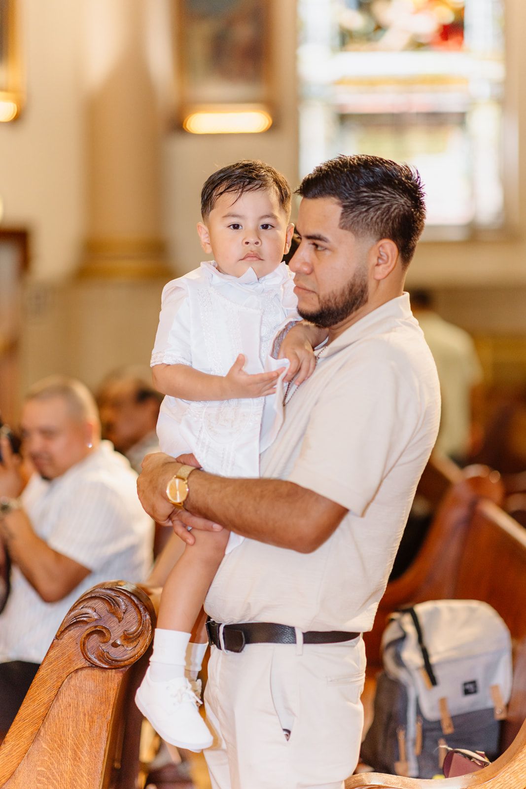 Man holding a baby dressed in white during a church ceremony, standing near wooden pews.
