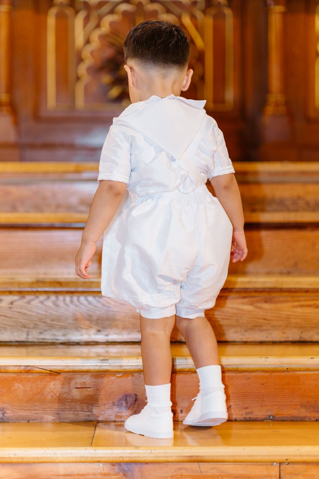 Toddler in white baptism outfit climbing wooden church steps, seen from behind, wearing white shoes and socks.