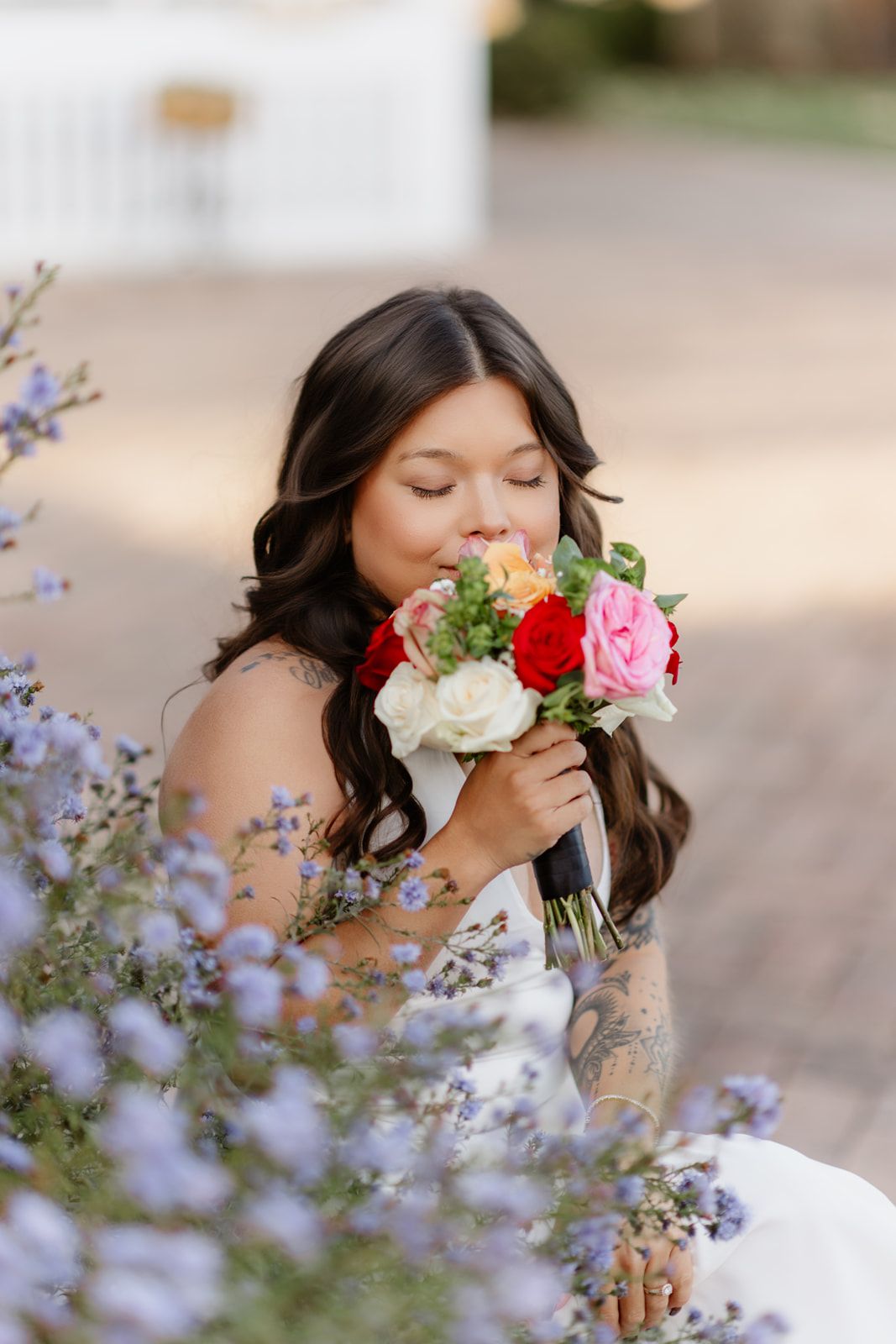 Tattooed bride in white dress smelling vibrant bridal bouquet with red and pink roses near purple flowers.