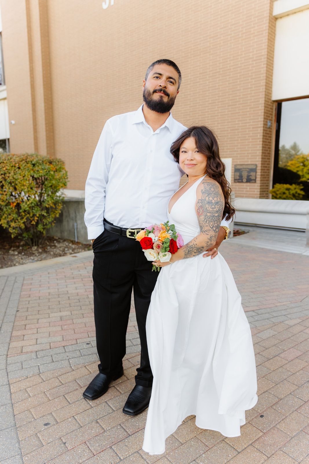 Newlywed couple poses outside courthouse, bride in white dress holding bouquet, groom in white shirt and black pants.